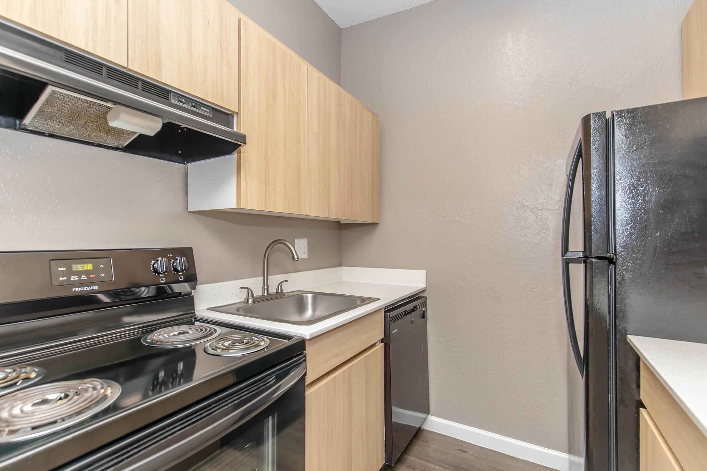 A modern kitchen featuring light wood cabinetry, black appliances including an oven, range, refrigerator, and dishwasher. The countertop is white, and there is a stainless steel sink. The walls are painted a neutral gray, creating a clean and contemporary look.