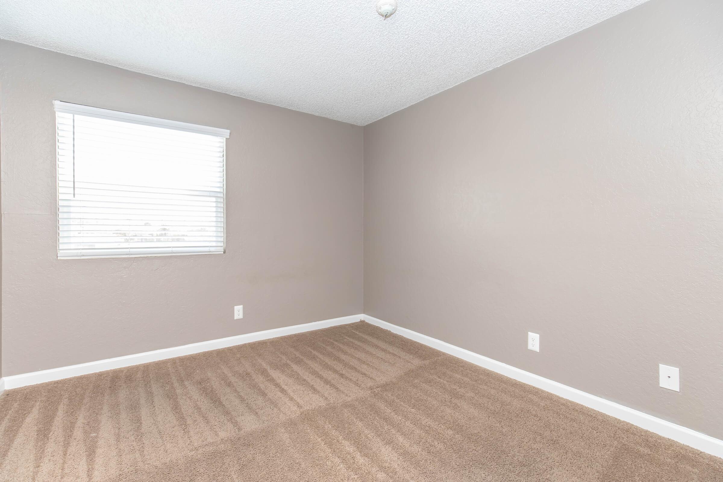 A vacant room with light brown walls and carpet. There is a window with white blinds allowing natural light in. The room appears clean and minimalistic, featuring a textured ceiling and no furniture, creating an open and empty space.