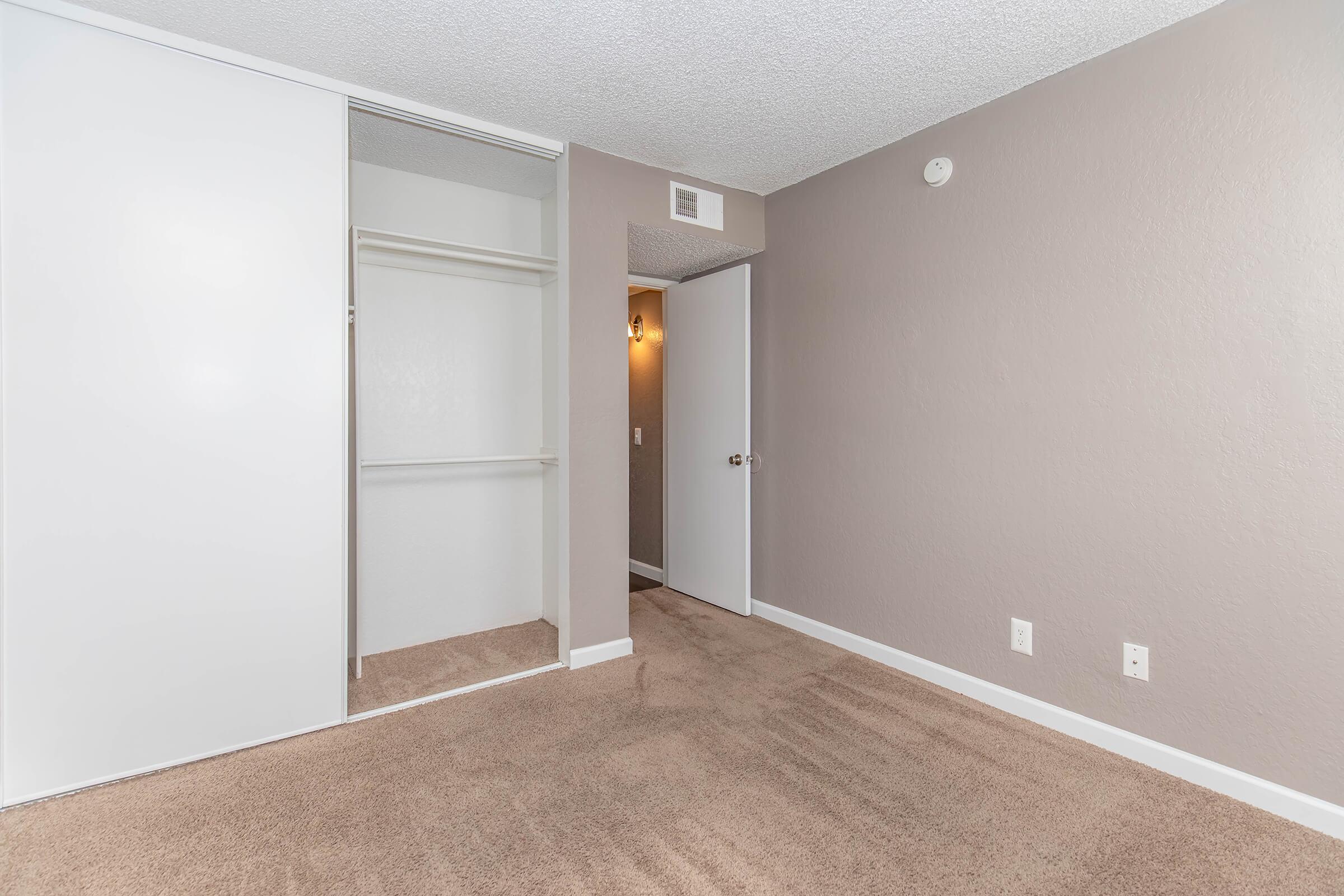 A clean, empty room with beige carpet. It features a closet with sliding doors on the left and a door leading to another area on the right. The walls are painted a soft, light color, and there is a light fixture visible on the wall. The space is bright and well-maintained, ideal for personalization.