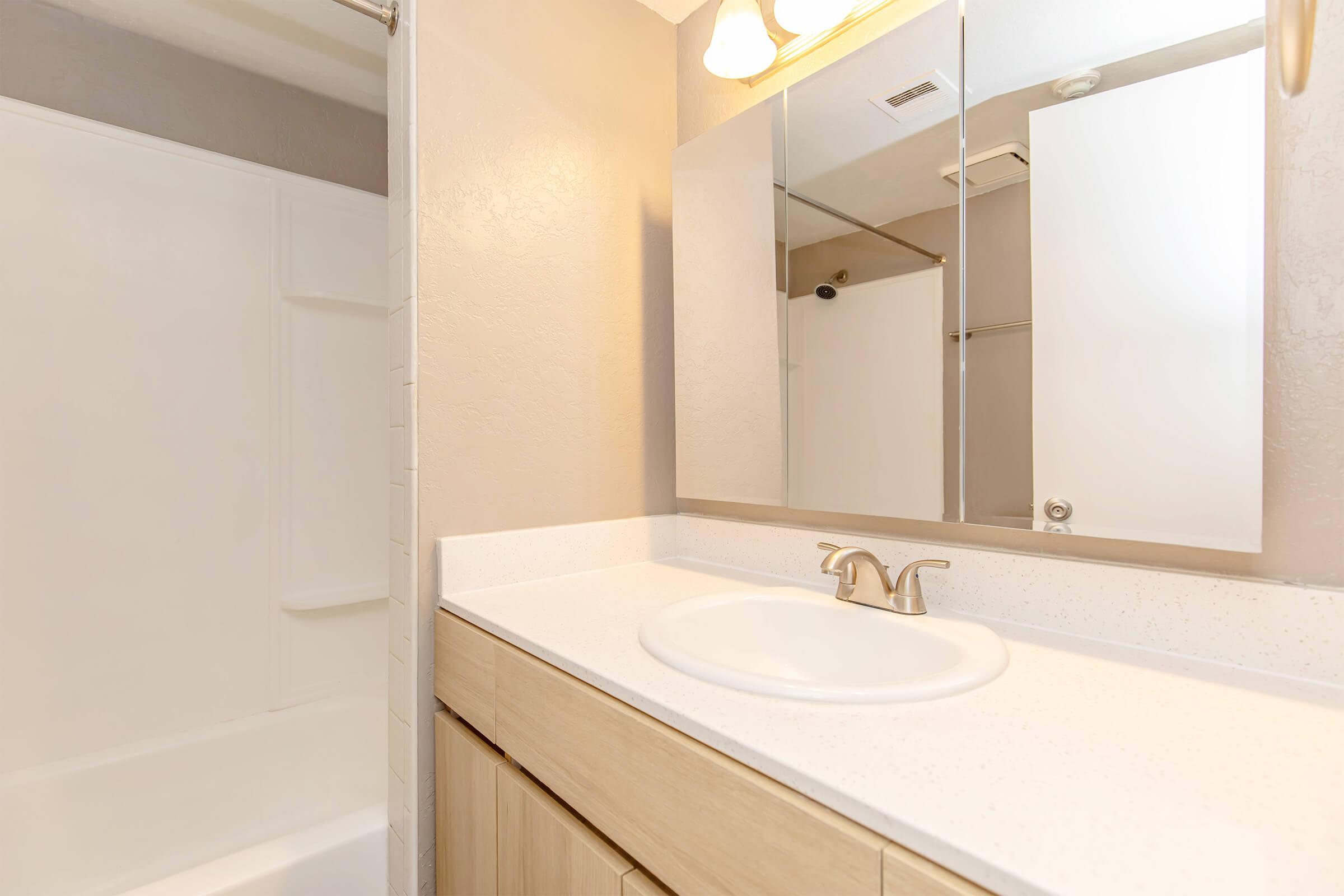 A bright bathroom featuring a white countertop with a round sink, light wooden cabinetry, a large wall mirror, and a shower area with a sliding glass door. The walls are a neutral tone, and overhead lighting provides a warm ambiance.