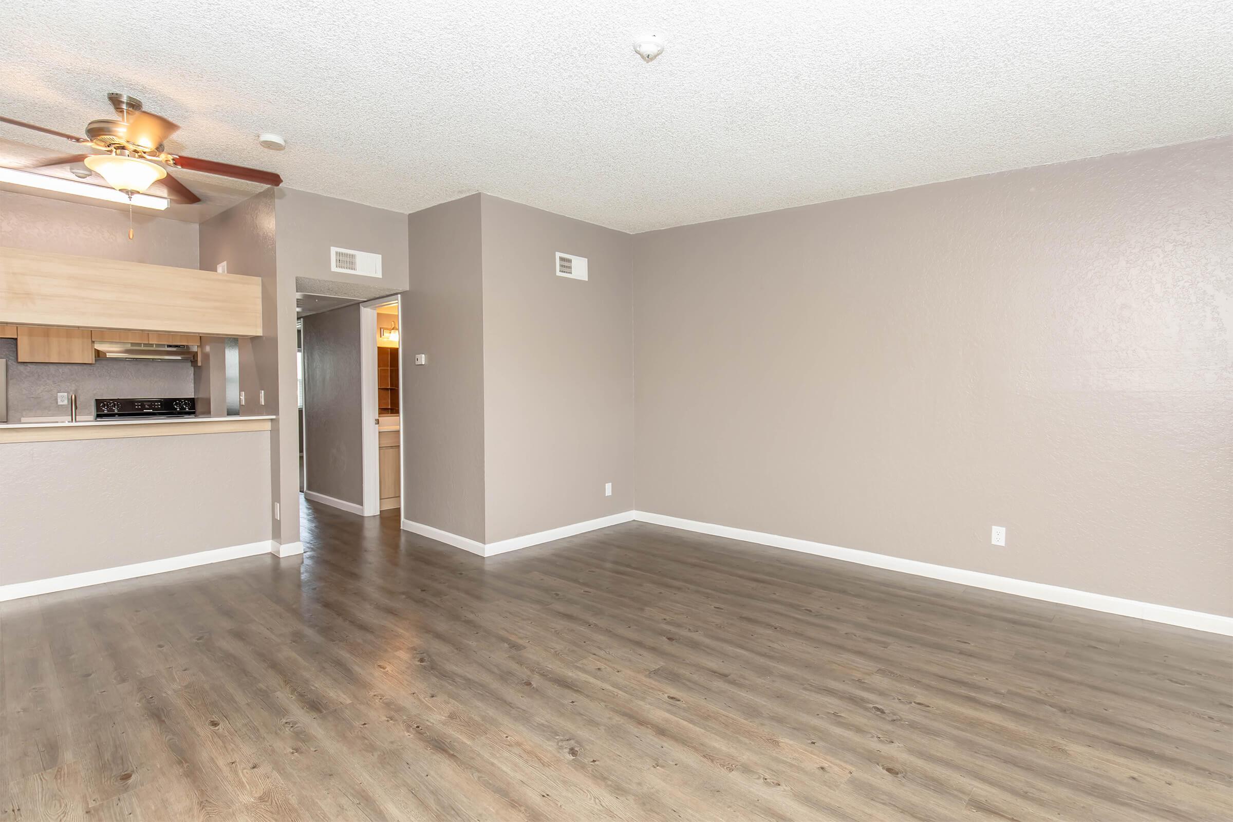 A spacious, empty room with light brown walls and hardwood flooring. A ceiling fan is visible, and there's an open kitchen area in the background. A doorway leads to another room, while natural light fills the space, creating a bright and airy atmosphere.
