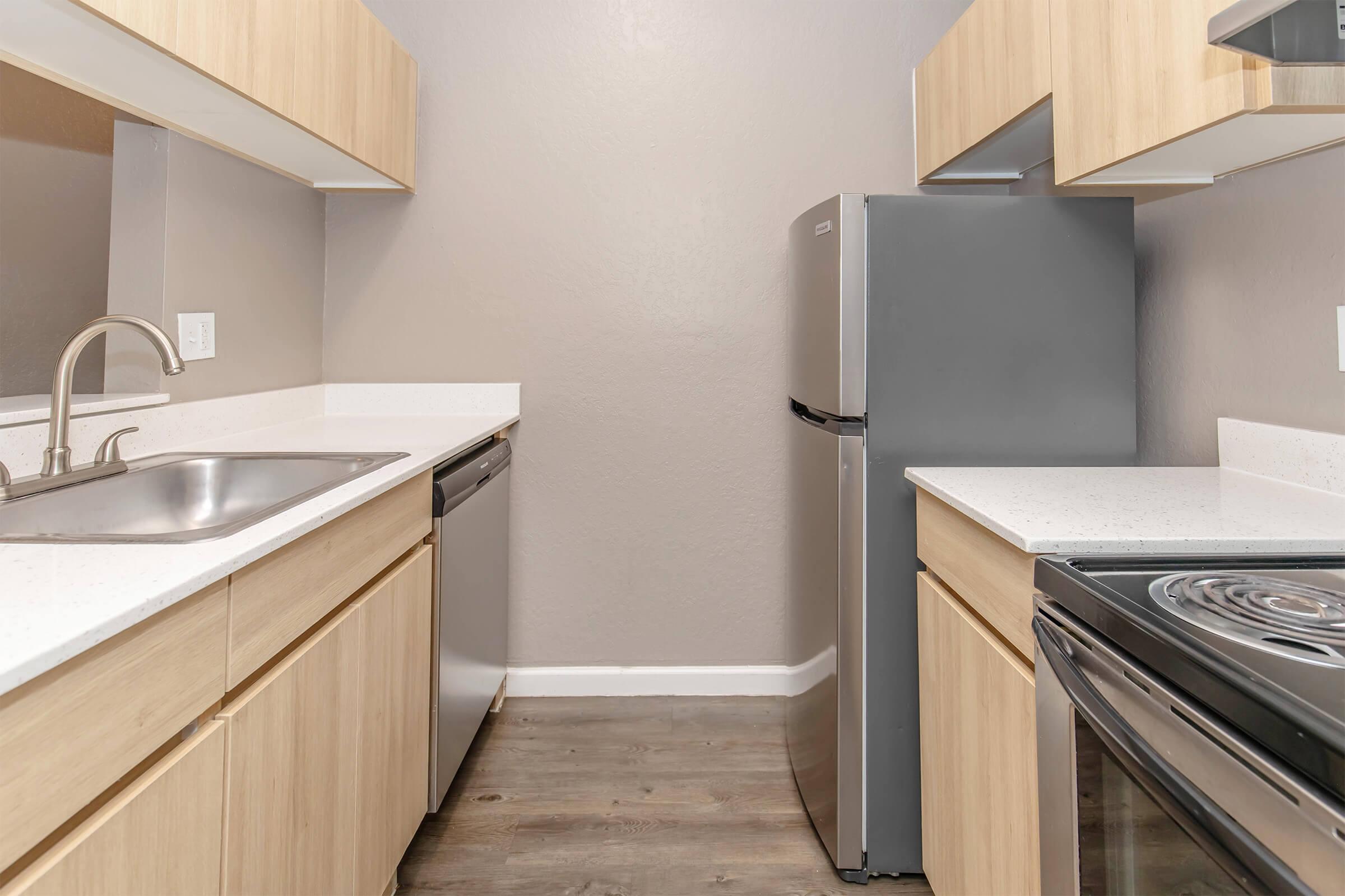 A modern kitchen featuring light wood cabinetry, a stainless steel refrigerator, a dishwasher, and a sink on one side, with a stove and oven across from it. The countertops are white, and the walls are painted a soft gray, creating a clean and contemporary look.
