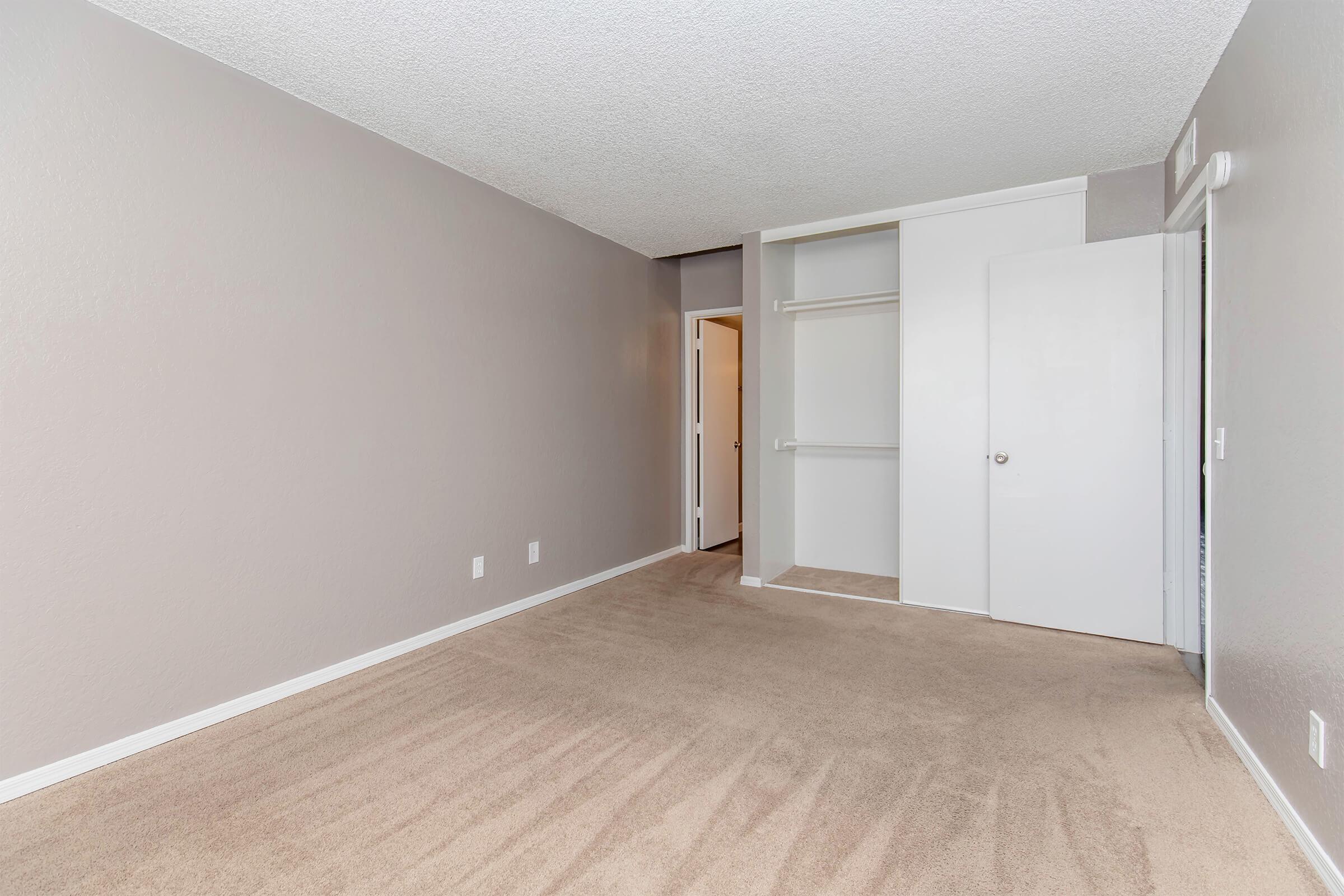 A vacant room with light gray walls and plush beige carpeting. There is a closet with sliding doors on the left side, and a white door leading to another space is visible on the right. The room is well-lit, creating a spacious and neutral environment suitable for various uses.