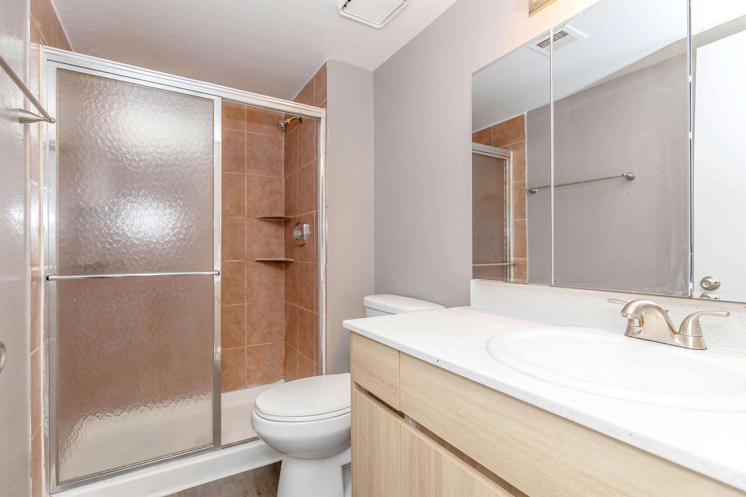 Modern bathroom featuring a glass-enclosed shower, beige tiled walls, and a vanity with a white countertop. A large mirror reflects the space, and a toilet is positioned next to the vanity. The overall design is clean and functional with neutral colors.