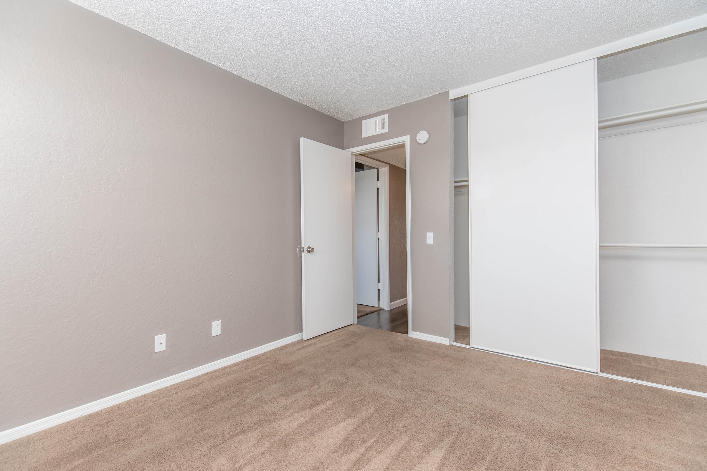 An empty room with beige carpet and light gray walls. A door leads to a hallway, and there's a closet with sliding white doors on one side. The lighting is bright and even, creating a clean and open atmosphere.