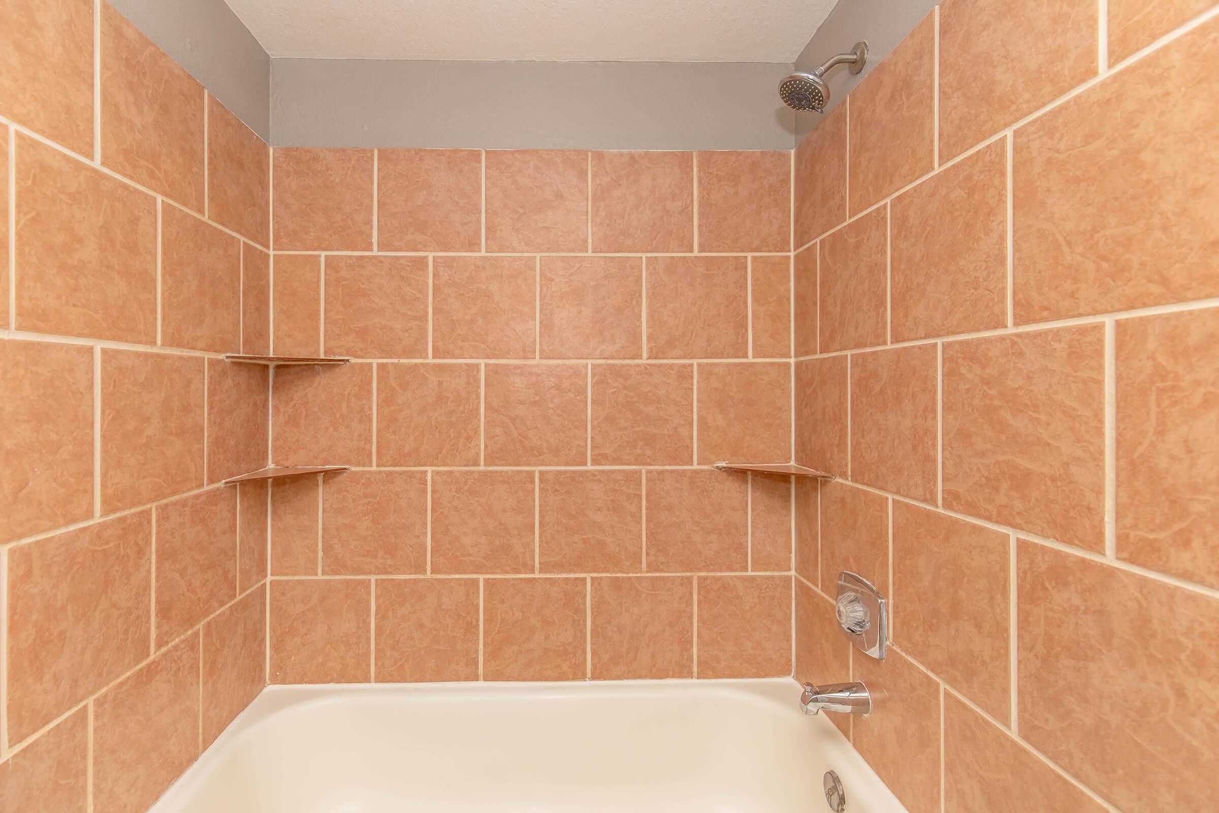 A bathroom shower area featuring beige tiled walls with a smooth finish, two built-in shelves for storage, and a white bathtub. The space is well-lit, with a showerhead mounted on the wall. The overall aesthetic is clean and minimalist.