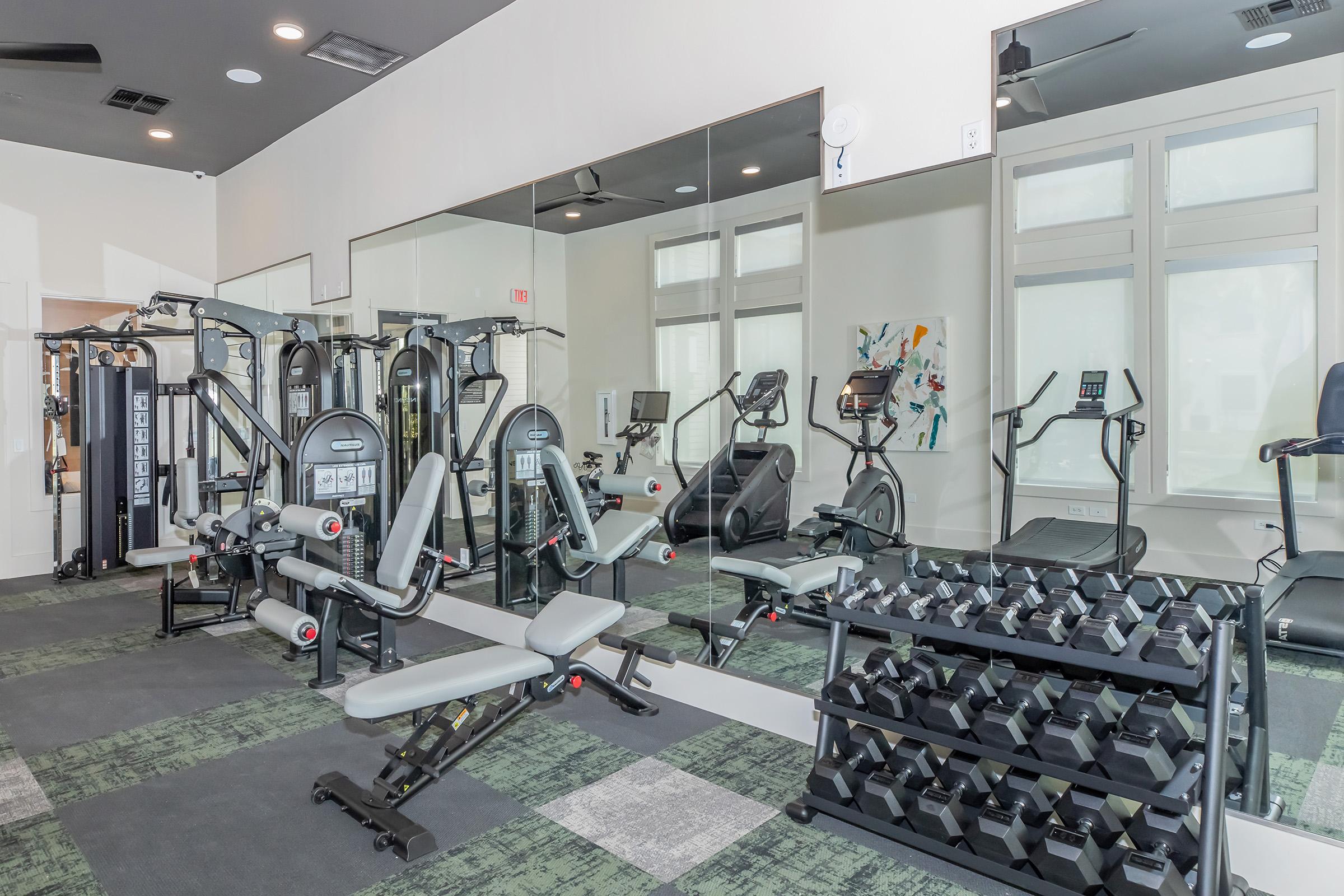 A modern gym interior featuring various exercise equipment including weight machines, treadmills, and free weights. Large mirrors reflect the space, and the flooring is a combination of carpet and tile. A bright, clean environment encourages fitness activities.