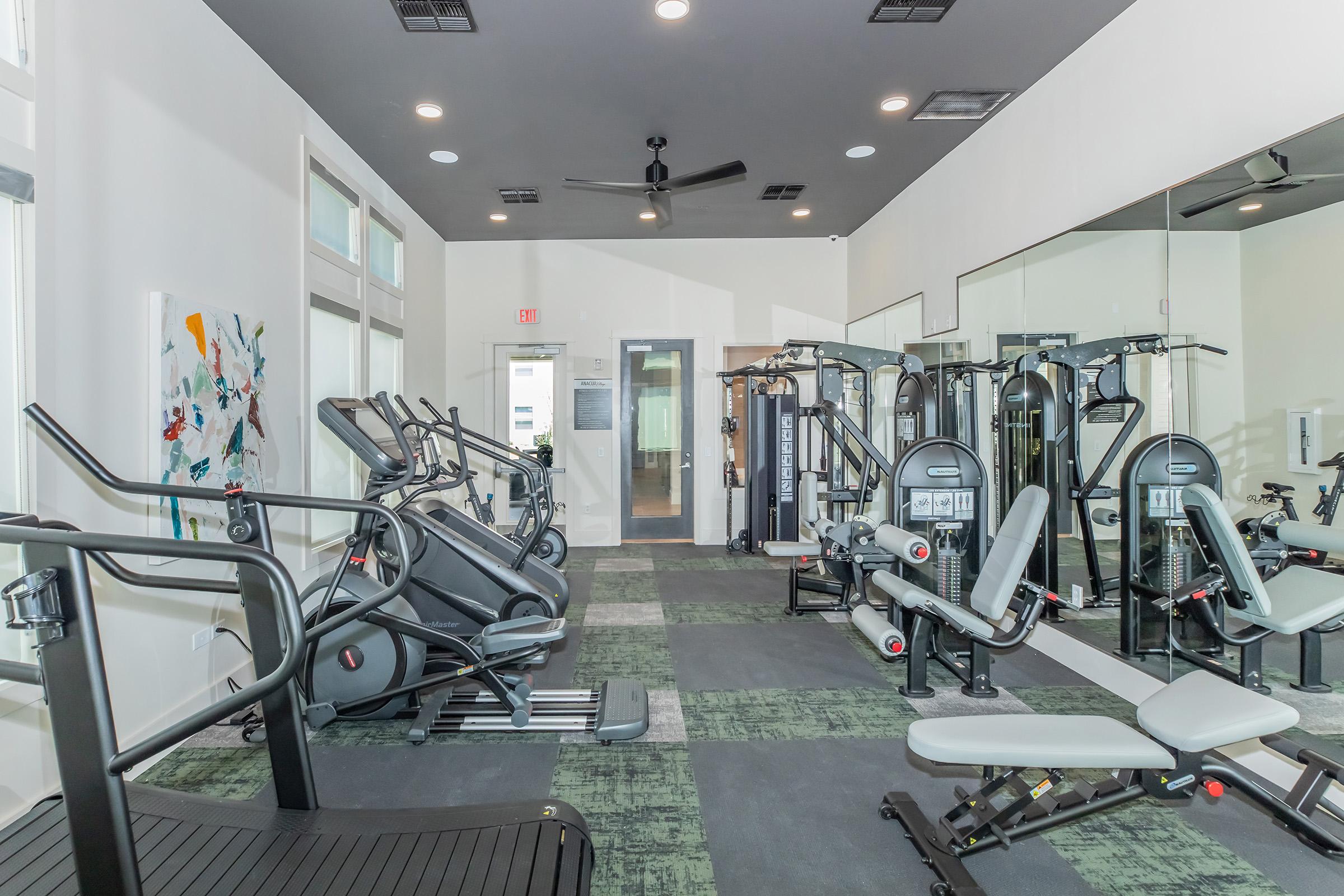 A modern gym interior featuring various exercise equipment, including cardio machines like treadmills and ellipticals, weight machines, and a mirrored wall. The space has a clean design with light-colored walls, green carpet, and ample natural light from large windows.