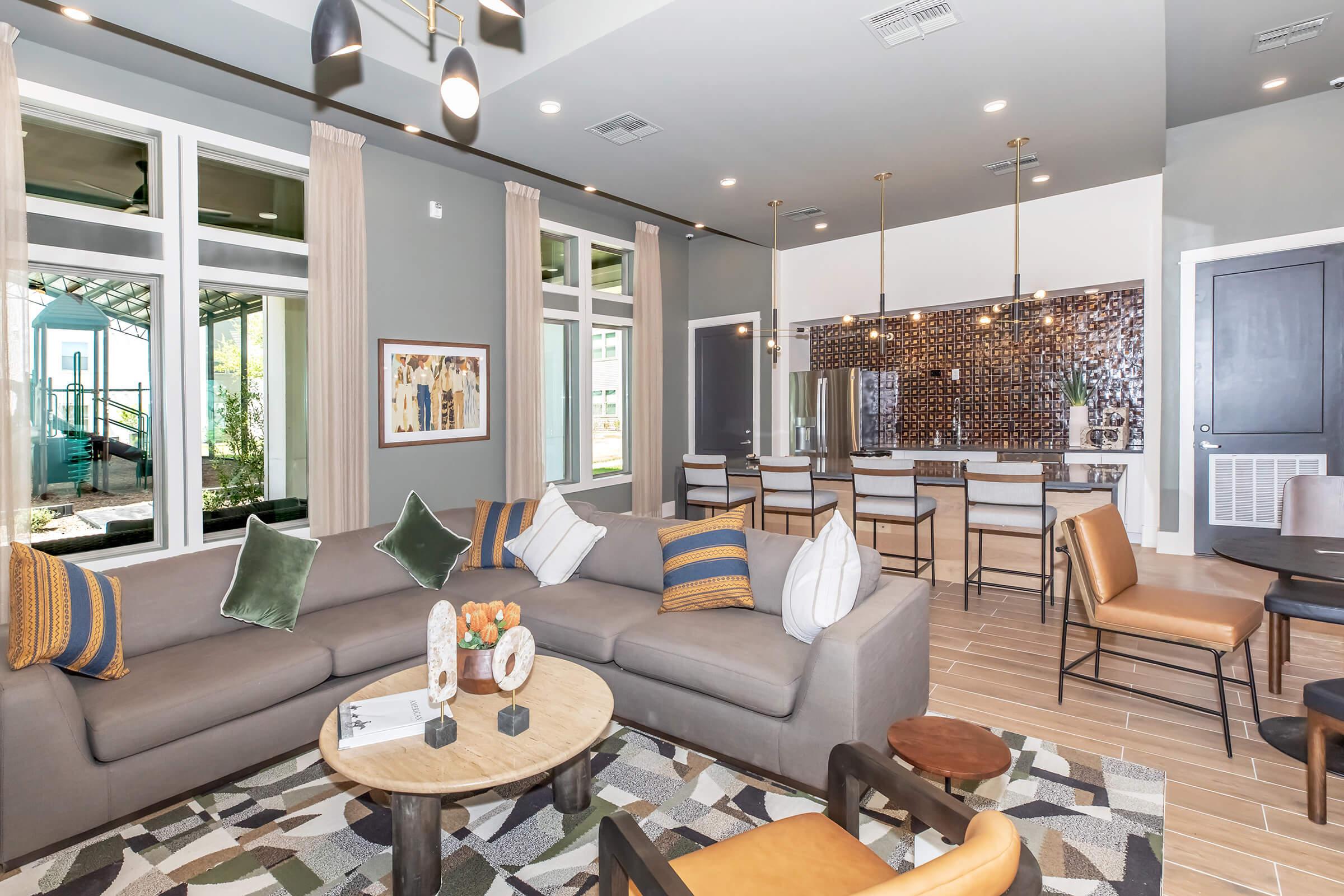 A modern living space featuring a sectional sofa with decorative cushions, a round coffee table, and a colorful area rug. Natural light streams in through large windows, and a sleek kitchen area is visible in the background, complete with bar seating and stylish pendant lights.