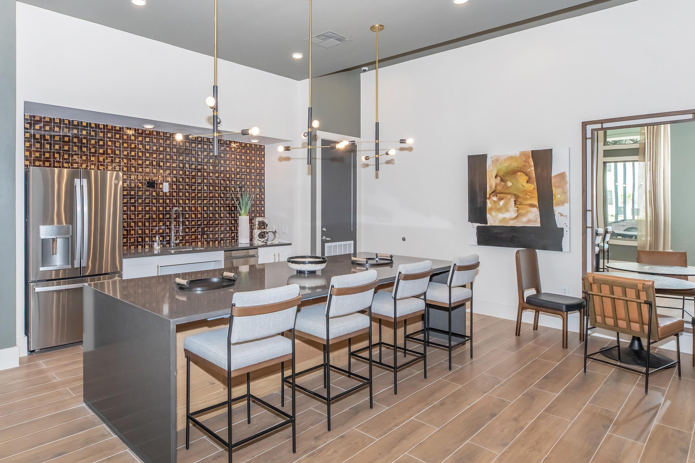 Modern kitchen featuring a sleek island with bar stools, stainless steel appliances, and a stylish backsplash. Natural light illuminates the space, complemented by contemporary lighting fixtures. A dining area with wood and metal furniture is visible, creating a warm and inviting atmosphere.