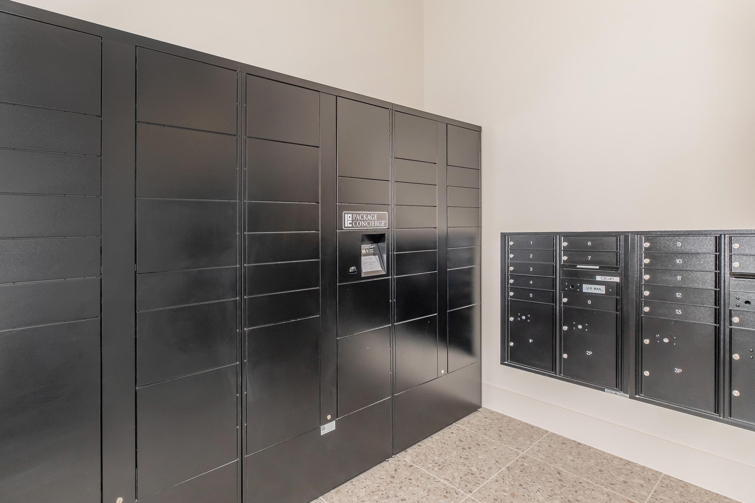 A modern package delivery area featuring black lockers for secure storage. The setup includes a central panel with a connectivity label, alongside smaller mailboxes. The floor is tiled, and the walls are simple and neutral in color, creating a functional, organized space for receiving parcels.