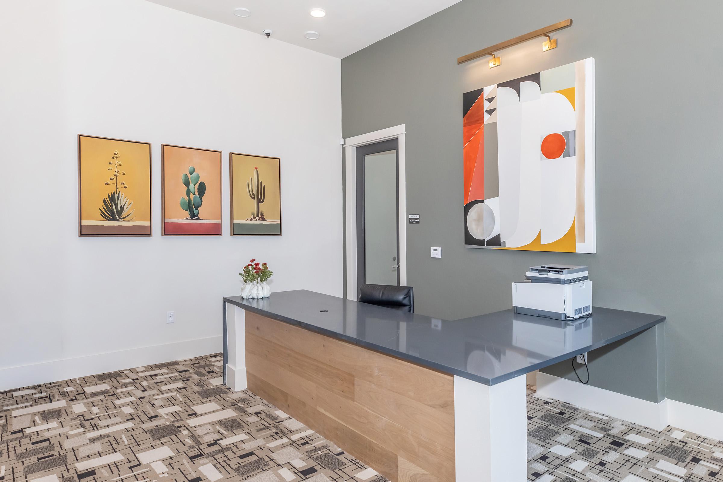 Modern reception area featuring a sleek wooden front desk, a printer, and three framed artworks depicting cacti on the wall. The background has a stylish gray paint, and there are decorative elements like a vase of flowers on the desk, with patterned flooring adding texture to the space.