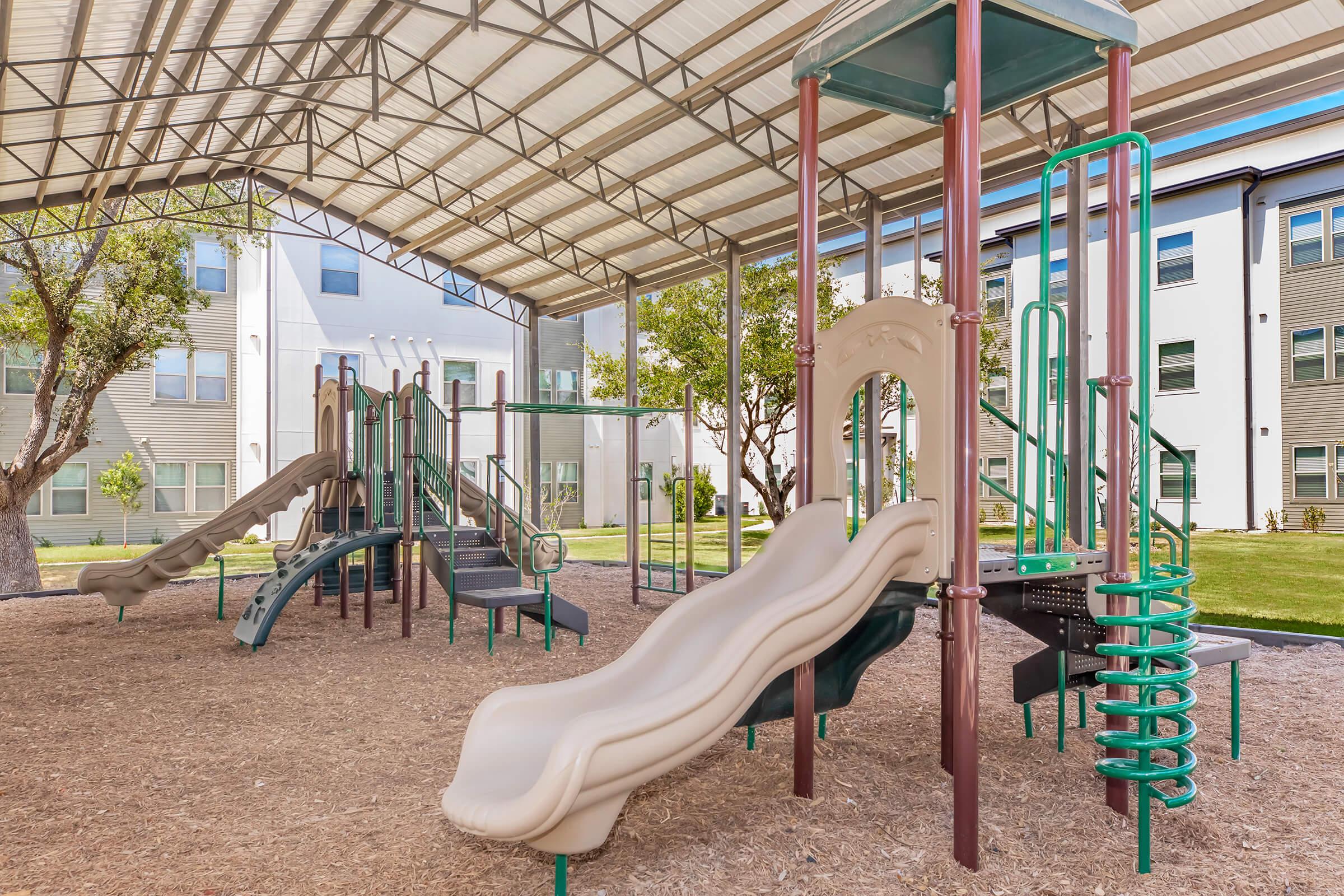 An outdoor playground featuring a covered play area with slides, climbing structures, and various play equipment, situated on a sandy surface. Surrounding the playground are grassy areas and nearby buildings, creating a vibrant recreational environment.