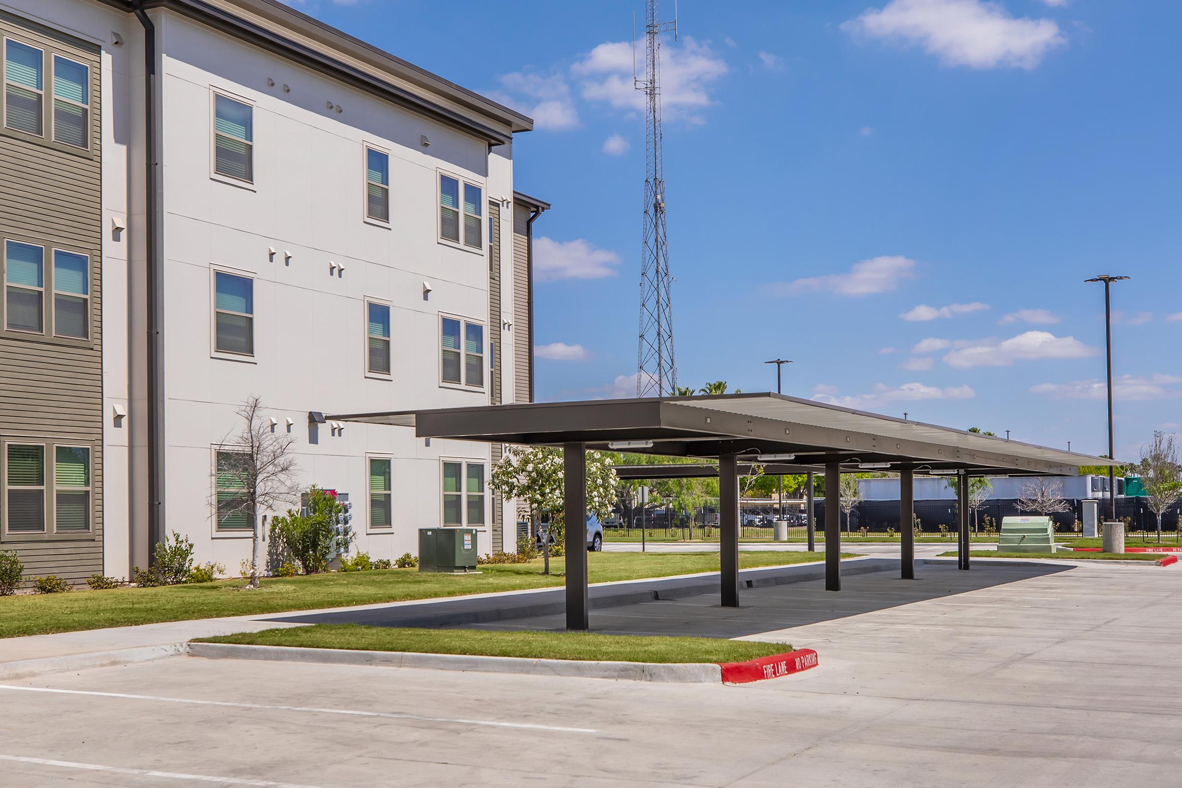 A modern building with multiple stories and a flat roof is adjacent to a spacious parking lot. There is a shaded parking structure with several spaces underneath, surrounded by green lawns and a few trees. The sky is blue, with scattered clouds, and a radio tower is visible in the background.