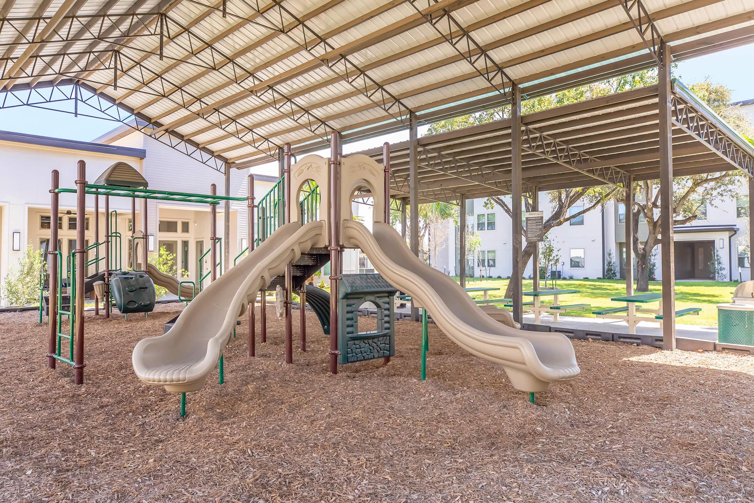 Playground structure with two tan slides under a large covered area. Surrounding the slides are climbing structures and a playhouse. The ground is covered with wood mulch, and green grass and trees are visible in the background. The setting appears bright and inviting for children.