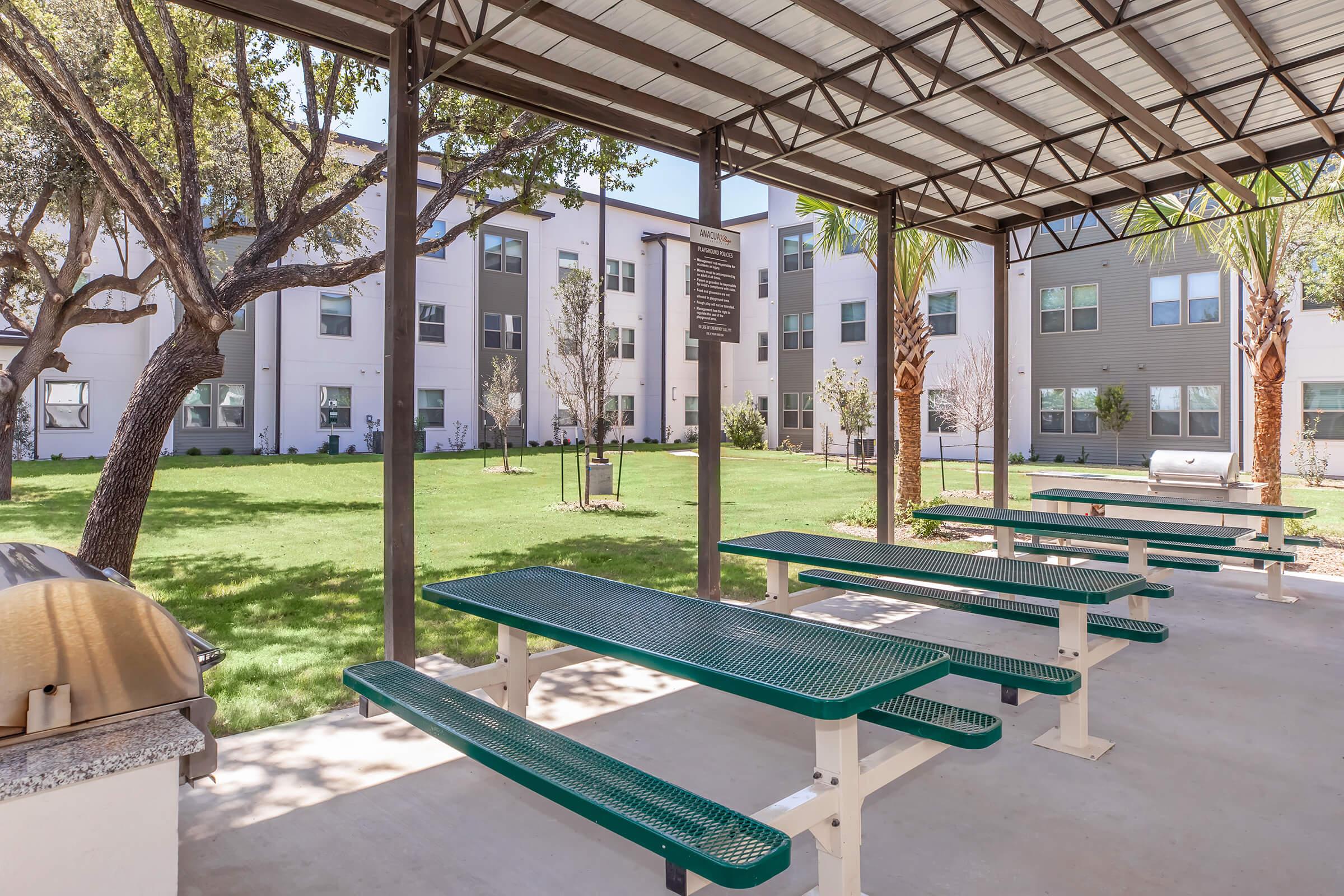 A shaded outdoor area featuring several green picnic tables under a metal canopy, overlooking a grassy yard with trees and a building in the background. The setting suggests a communal space suitable for gatherings or relaxation.