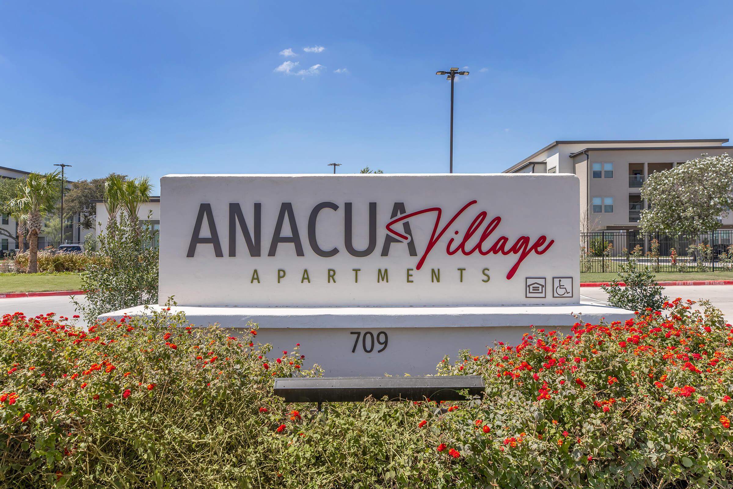 Sign for Anacua Village Apartments, prominently displaying the name in red cursive letters. The sign features decorative landscaping with vibrant red flowers and greenery. In the background, modern apartment buildings can be seen under a clear blue sky. The location number, 709, is also included on the sign.