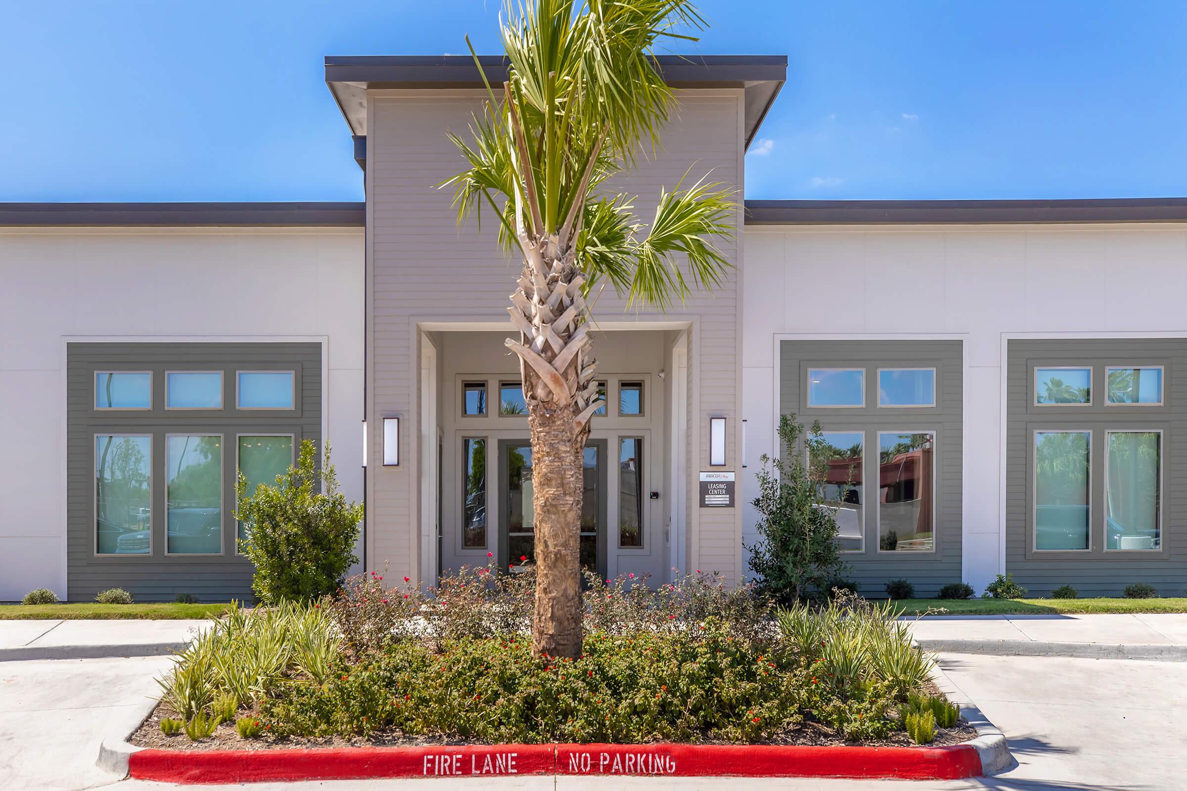 Modern building entrance with large windows, palm trees, and landscaping. The foreground features a circular flower bed with various plants, while a red curb with a "No Parking" sign is visible. The facade has a light color scheme and a small sign near the entrance. Clear blue sky in the background.