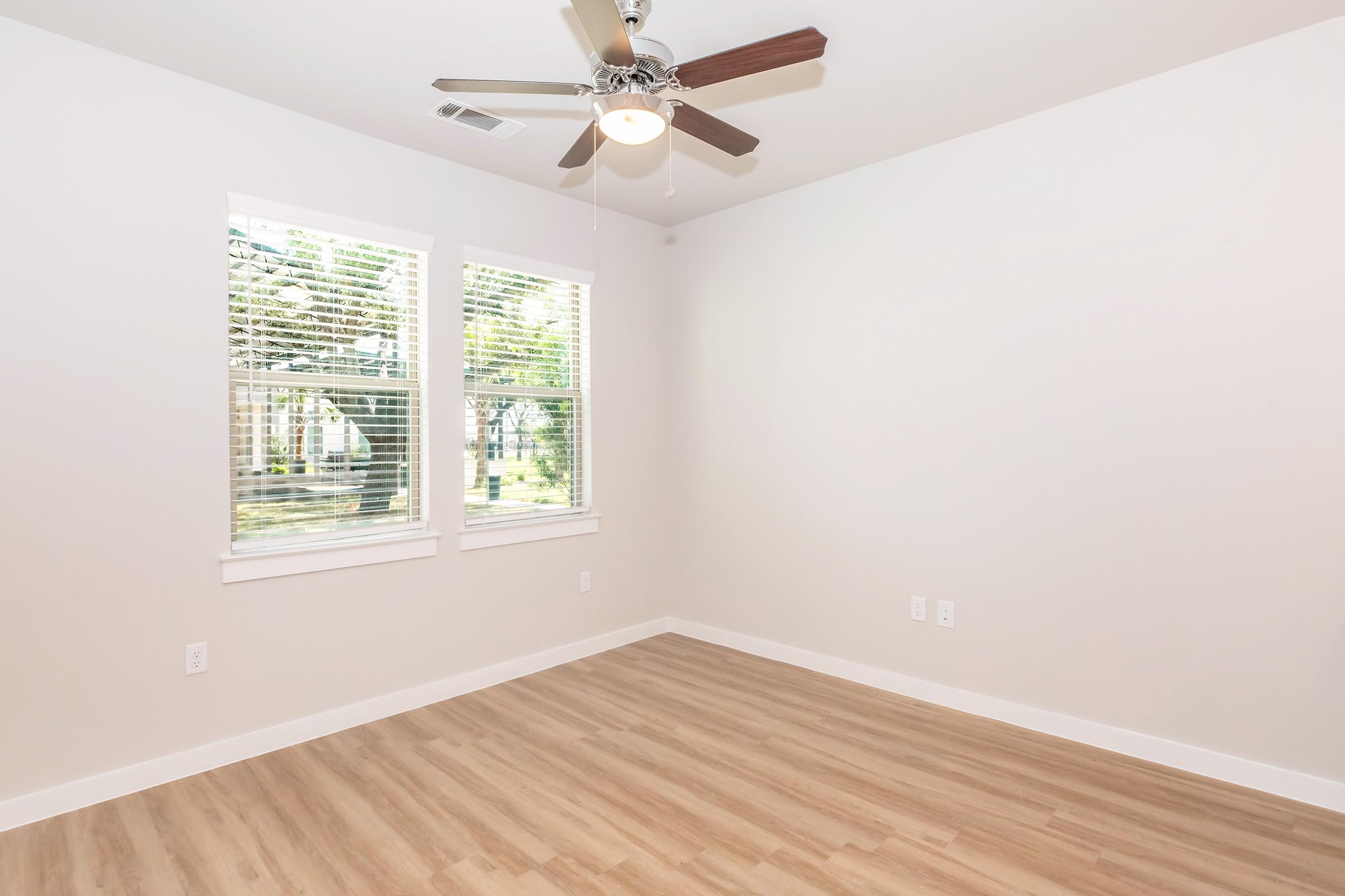 Bright and empty room with light wood flooring, two windows allowing natural light, and a ceiling fan. The walls are painted in a neutral color, creating a spacious and airy feel.