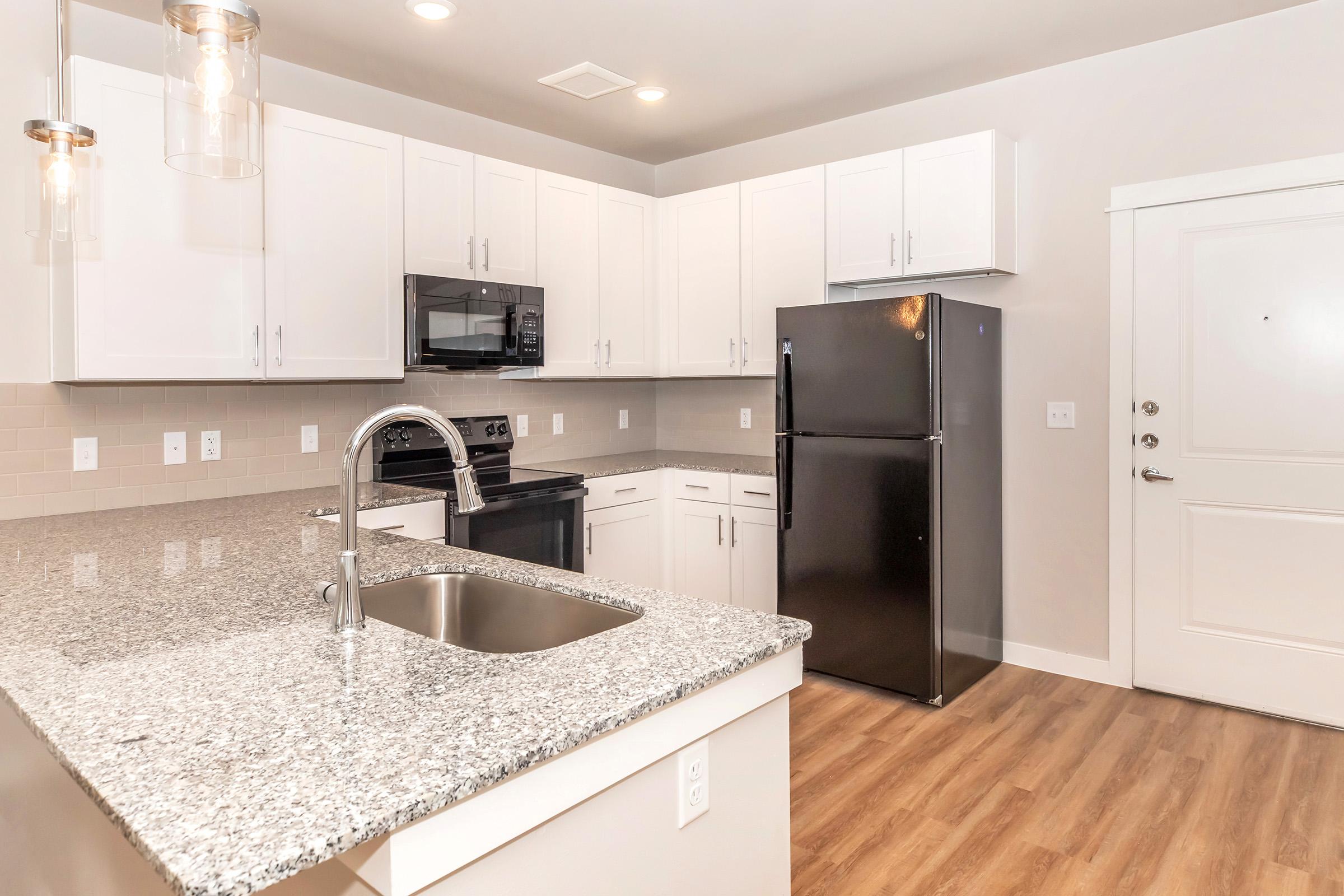 Modern kitchen featuring white cabinetry, a gray granite countertop, stainless steel appliances including a microwave and black refrigerator, and a door leading outside. The space has warm wooden flooring and contemporary pendant lighting.