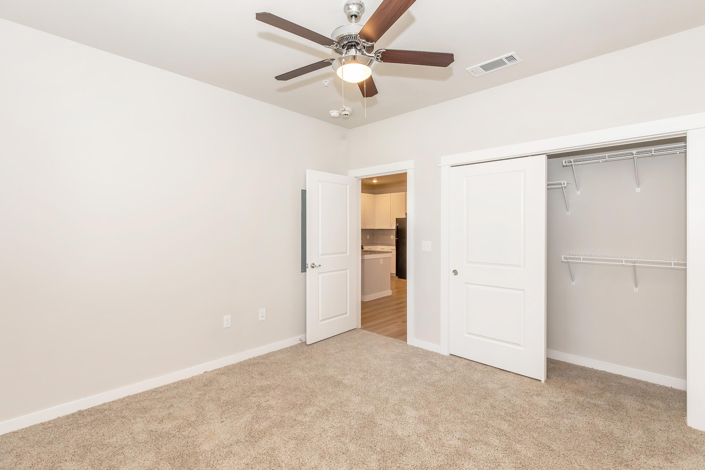A spacious bedroom featuring light-colored walls and plush carpet. It includes a ceiling fan with wooden blades, a door leading to another room, and a closet with sliding doors. Natural light highlights the openness of the space, creating a welcoming atmosphere.