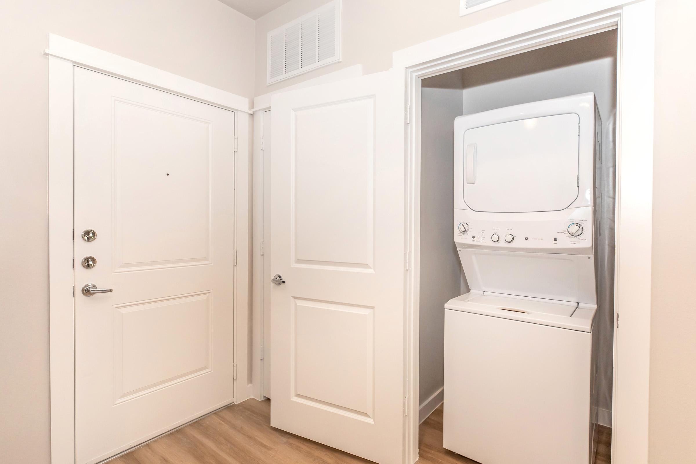 A tidy laundry area featuring a stacked washer and dryer unit, situated within a clean, well-lit hallway next to a white front door. The walls are painted in neutral tones, and the floor has light-colored wood-like laminate.