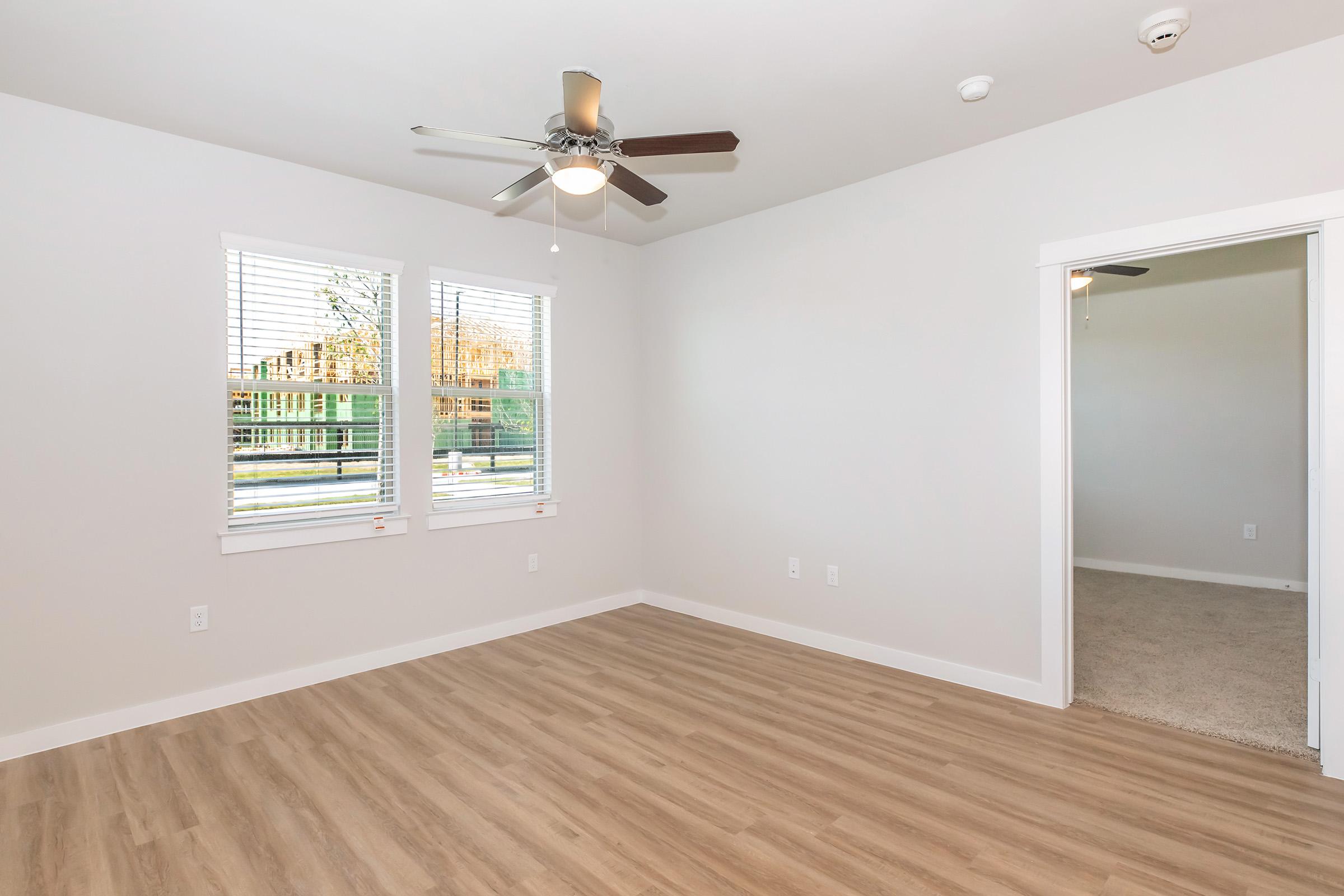A clean and empty room featuring light-colored walls, a ceiling fan, and large windows providing natural light. The floor is covered with light wood-like laminate, and there is a doorway leading to another room partially visible on the right.