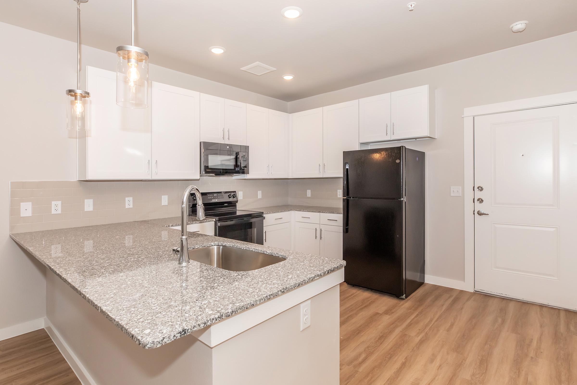 Modern kitchen featuring white cabinets, stainless steel appliances including a microwave and oven, a black refrigerator, and a large countertop with a sink. The flooring is a light wood finish, and there are pendant lights hanging above the island. A door leads to the entrance.