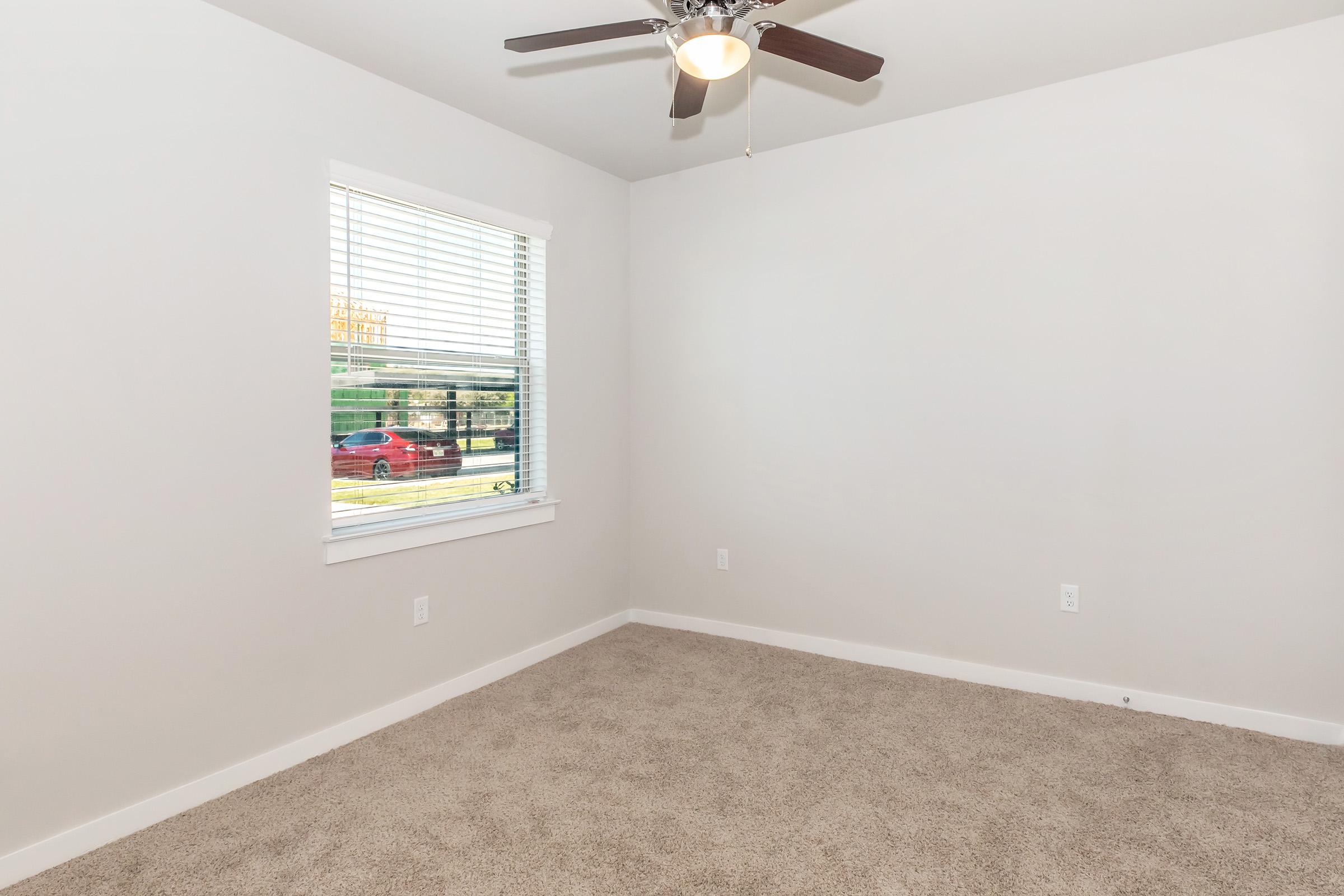 Empty room with light beige carpet and a light gray wall. A single window with white blinds lets in natural light, revealing a glimpse of greenery and a red car outside. A ceiling fan with wooden blades hangs from the ceiling, adding to the clean and simple decor.