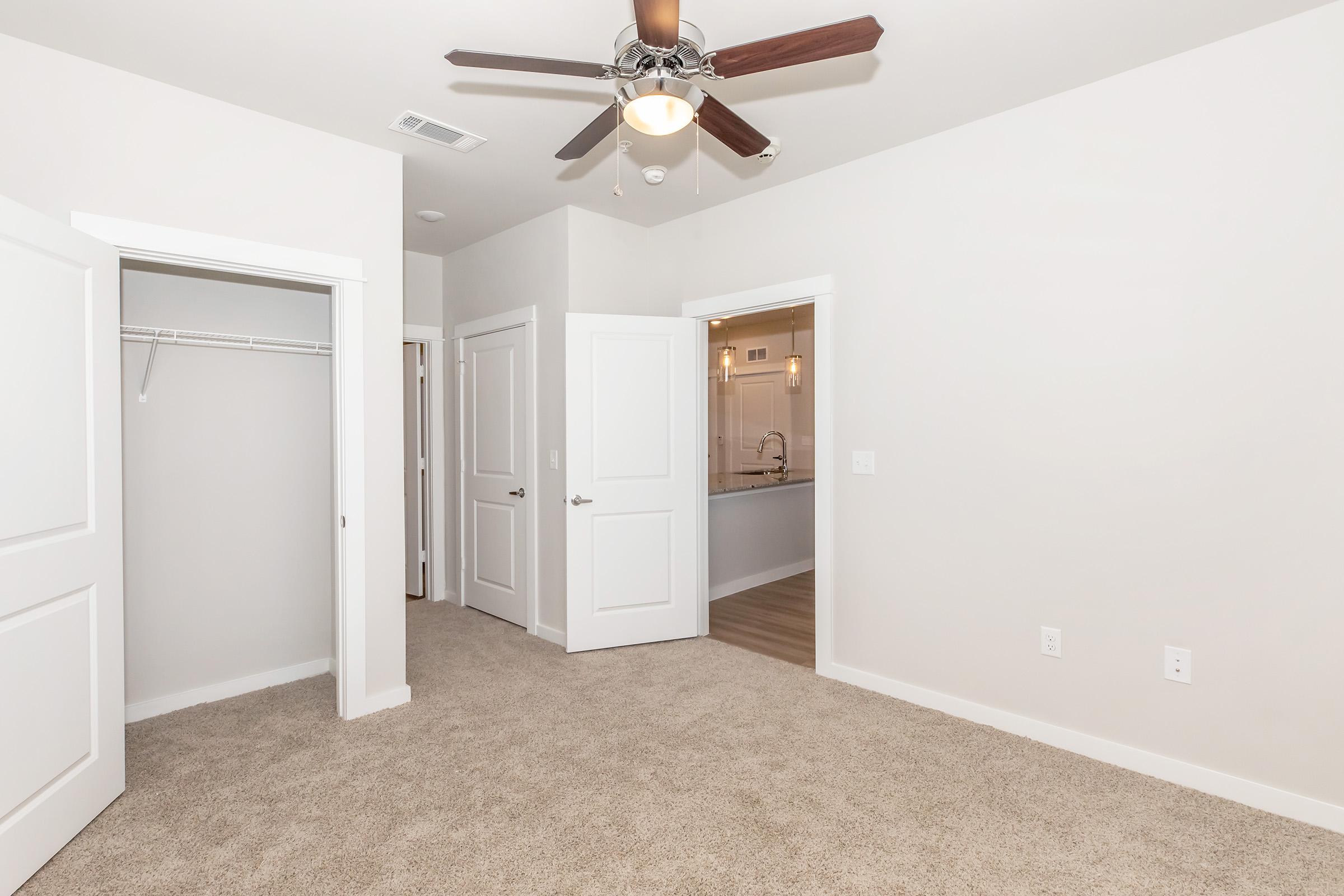 A clean, well-lit room with beige carpet and light gray walls. The space features a ceiling fan, two opened doors leading to a closet and another room, and is designed with a contemporary aesthetic. A glimpse of a bathroom can be seen through one of the doorways.
