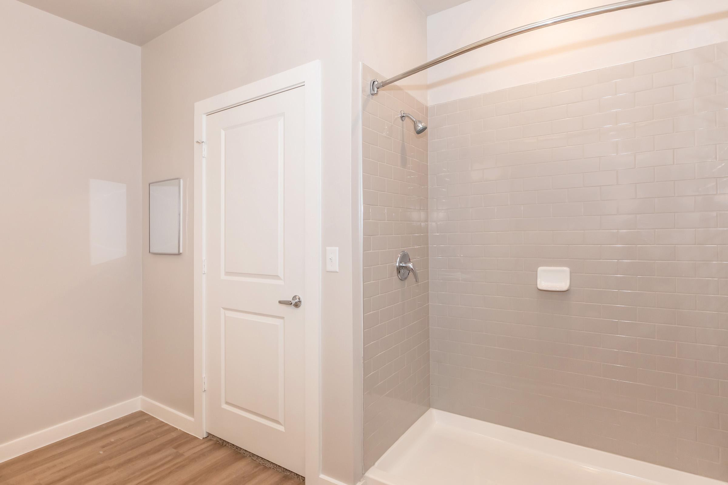 A clean and modern bathroom featuring a shower stall with tiled walls, a glass door, and a white base. The walls are painted a light color, and there's a wooden floor. A closed white door is visible, along with a panel on the wall, suggesting a minimalist design.