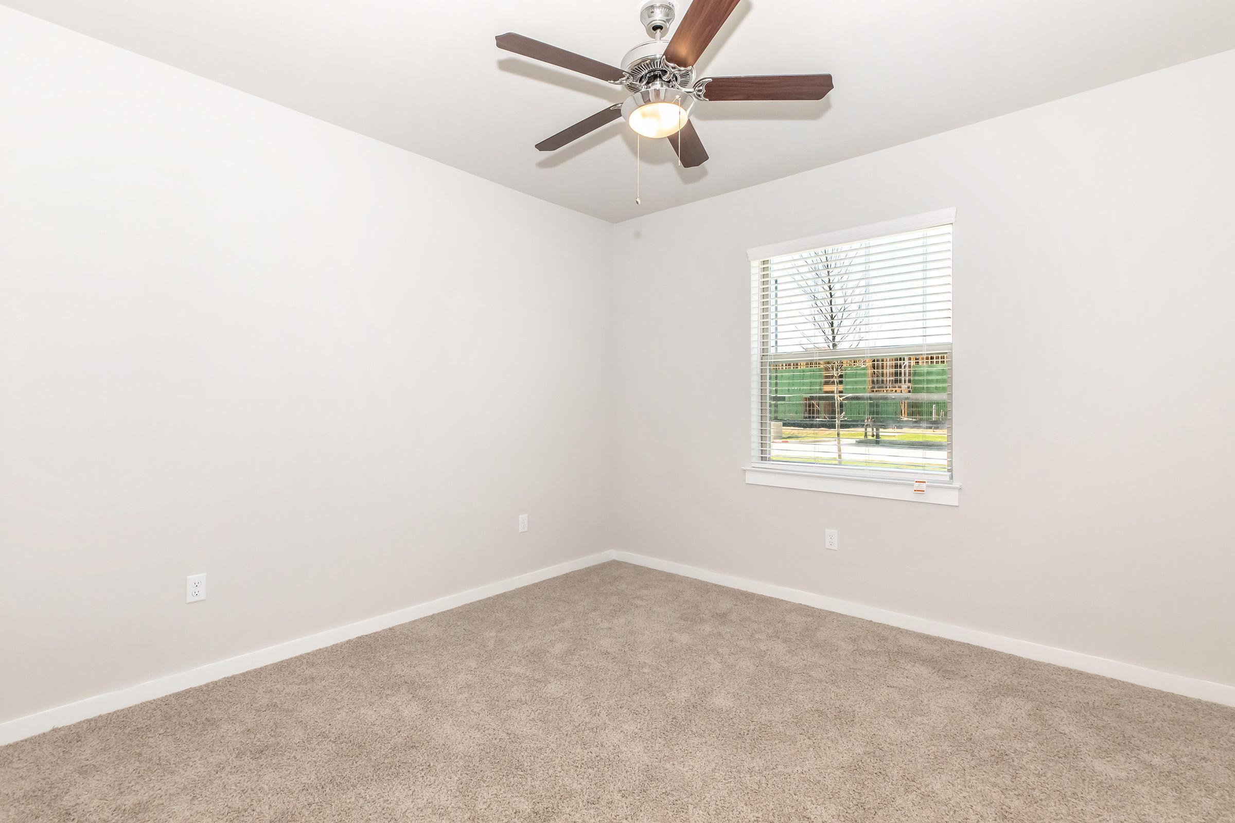A vacant room featuring light gray walls, a ceiling fan with wooden blades, and a large window with white blinds. The floor is covered in beige carpet, creating a spacious and airy atmosphere. Natural light streams in through the window, highlighting the simplicity of the space.