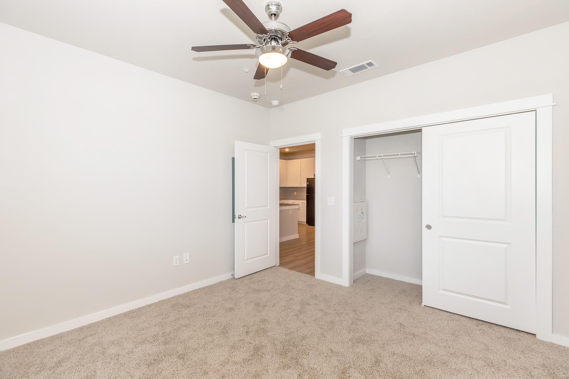 A light-colored room with plush carpet, featuring a ceiling fan and two doors. One door leads to a closet with shelves, while the other opens to a bright area, possibly a kitchen. The walls are painted white, and the overall space appears clean and modern. Natural light may be coming from the adjacent room.
