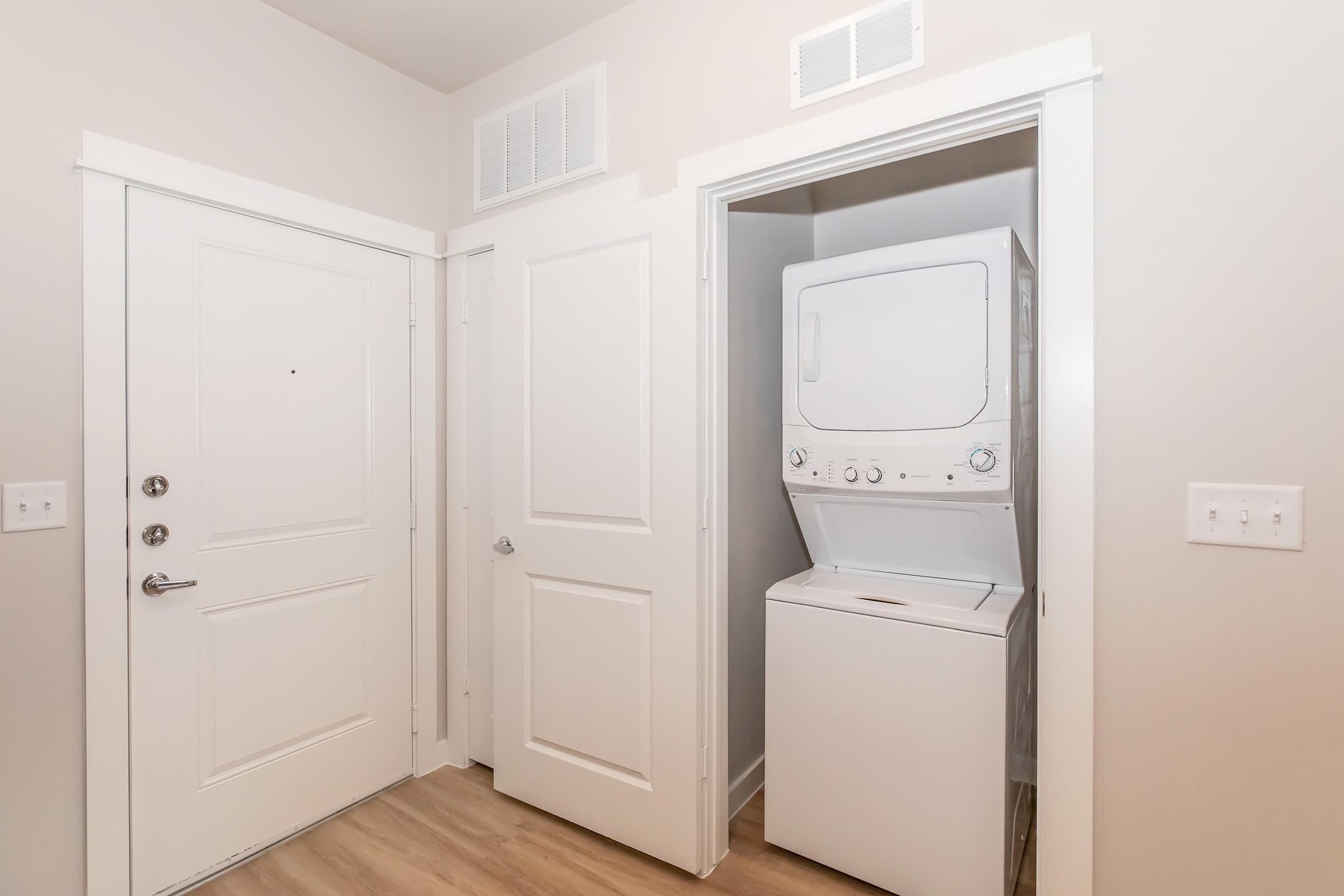 A small laundry area featuring a stacked washer and dryer unit. The room has a light-colored wall and a doorway leading to the outside, with a white door and adjacent closet doors. The flooring is a light wood finish, creating a clean and modern look.