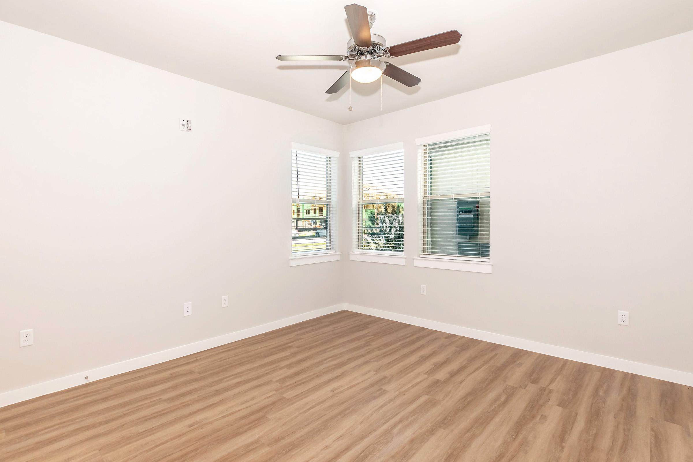 Empty room with light-colored walls and a ceiling fan. Three windows provide natural light, and the floor is finished with light wood laminate. The space appears clean and newly renovated, suitable for various uses like a living area or office.