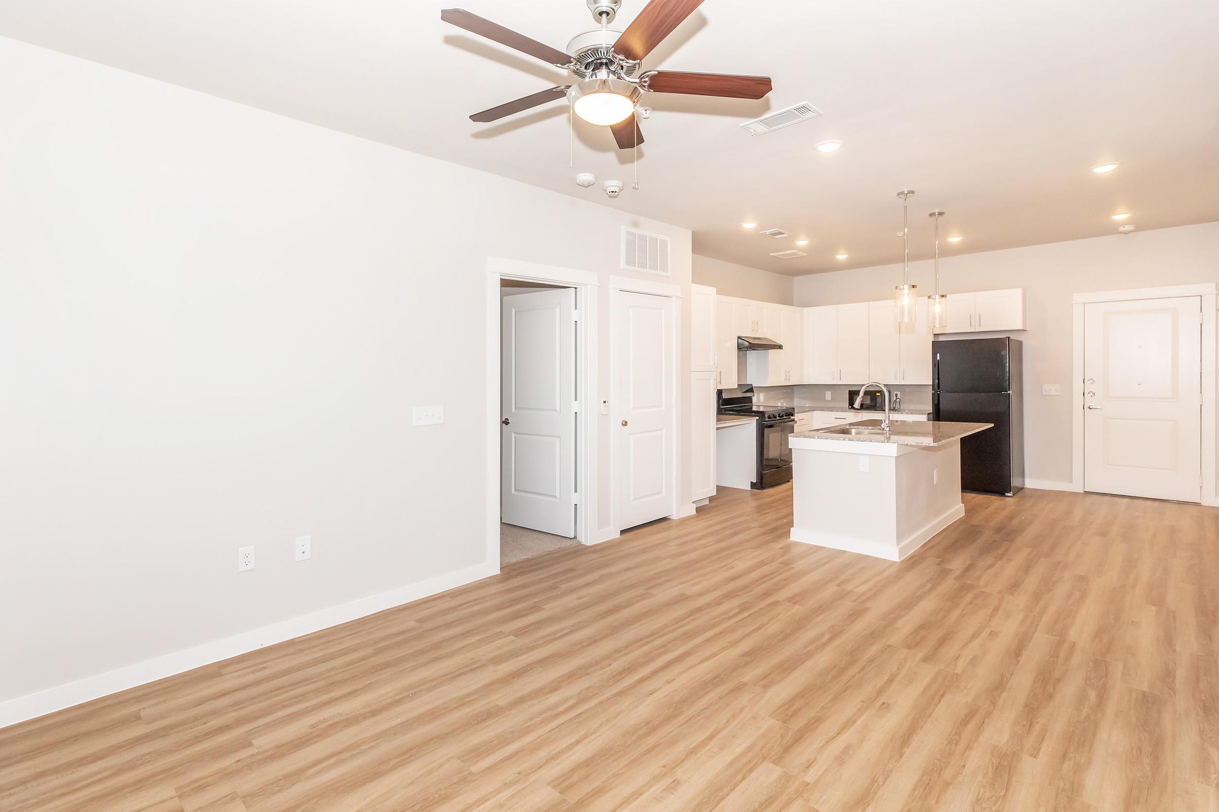 A modern interior of an apartment featuring a spacious living area with light wood flooring, a ceiling fan, and an open kitchen equipped with stainless steel appliances, white cabinetry, and a countertop. The room has neutral-colored walls and an inviting atmosphere.