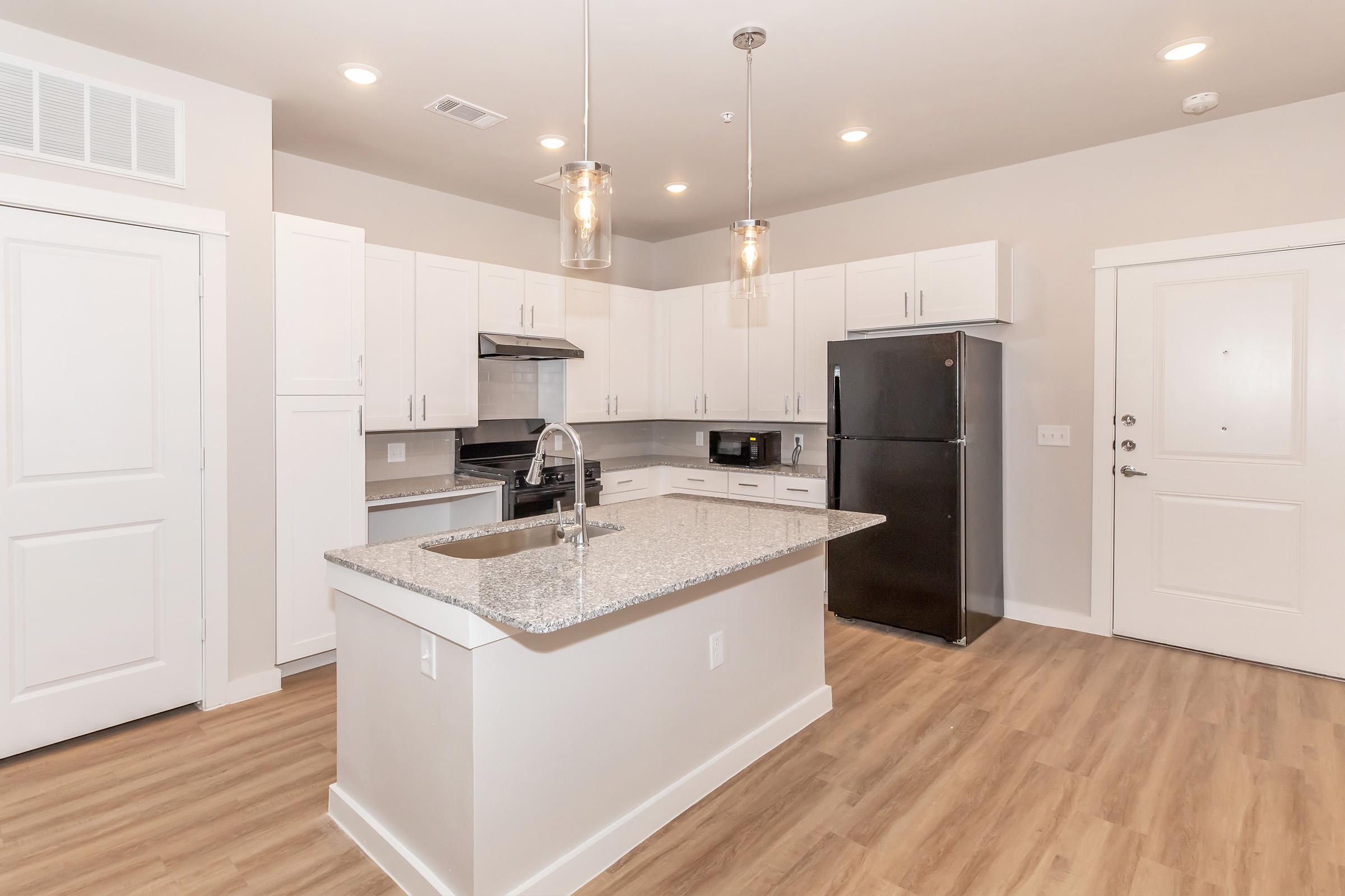 Modern kitchen featuring a spacious island with a granite countertop, stainless steel appliances, white cabinets, and pendant lighting. The open layout includes a door leading to the hallway and a black refrigerator. Warm wooden floors enhance the contemporary design.