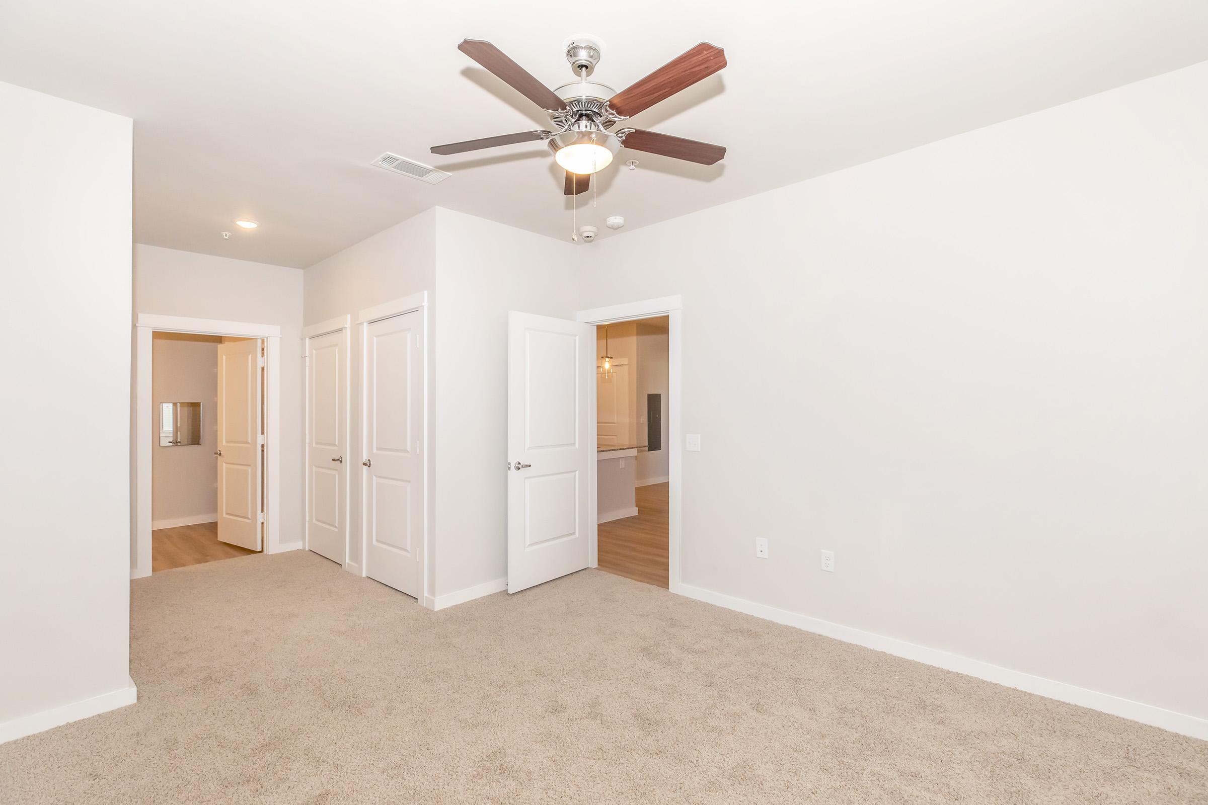 A spacious room featuring light beige carpet flooring, a ceiling fan with wooden blades, and multiple doors leading to adjacent areas. The walls are painted in a soft neutral tone, creating a bright and airy atmosphere. Natural light can be seen coming from one of the doorways.