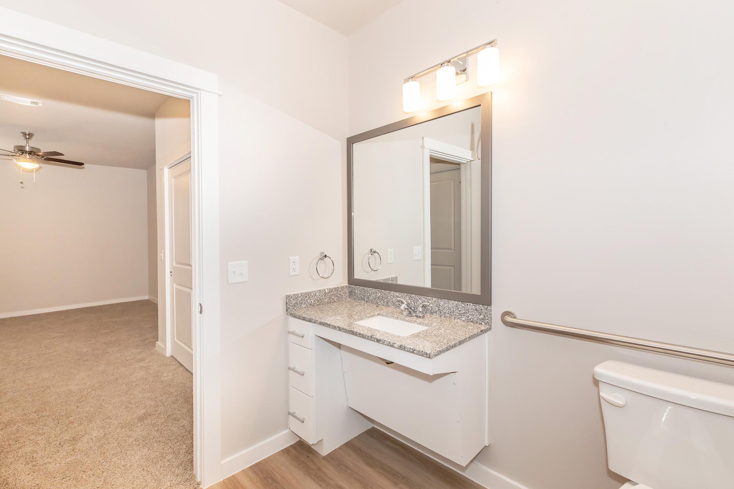 A modern bathroom featuring a vanity with a granite countertop and mirror, a toilet, and a towel rack. The walls are painted light, and warm lighting fixtures are above the mirror. There is a doorway leading to another room with carpet, suggesting a spacious layout.