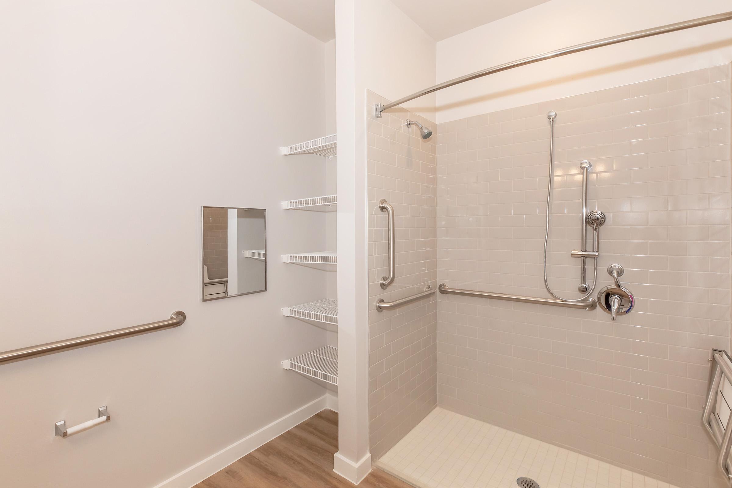A modern, accessible bathroom with a spacious walk-in shower. Features include grab bars for support, a handheld showerhead, and built-in shelves for toiletries. The walls are tiled in light colors, and there is a large mirror visible. The flooring is light wood, creating a clean and contemporary aesthetic.