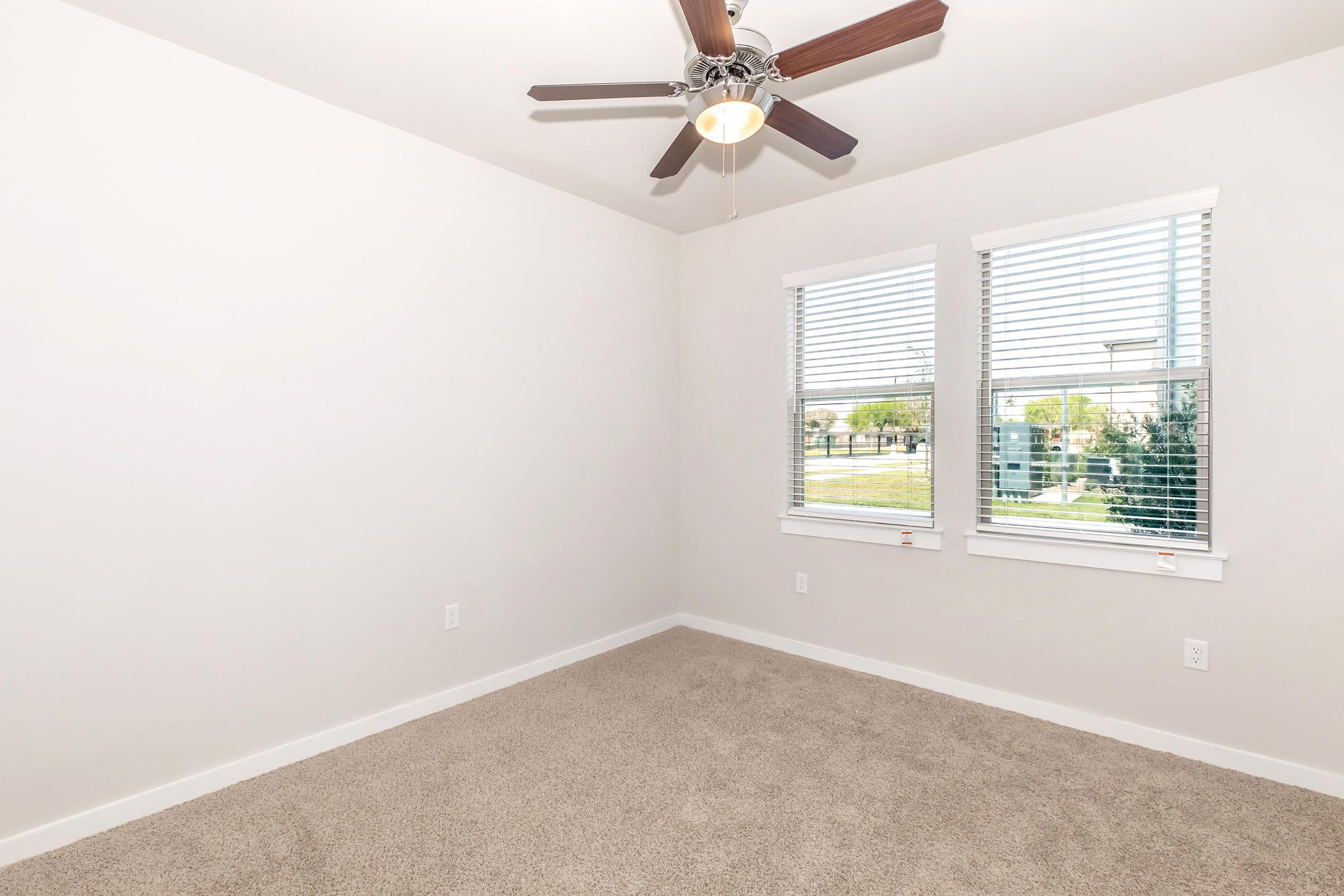 Empty room with light-colored walls and a ceiling fan. Two windows with blinds provide natural light, and the floor is covered with beige carpet. The view outside features greenery in the distance.