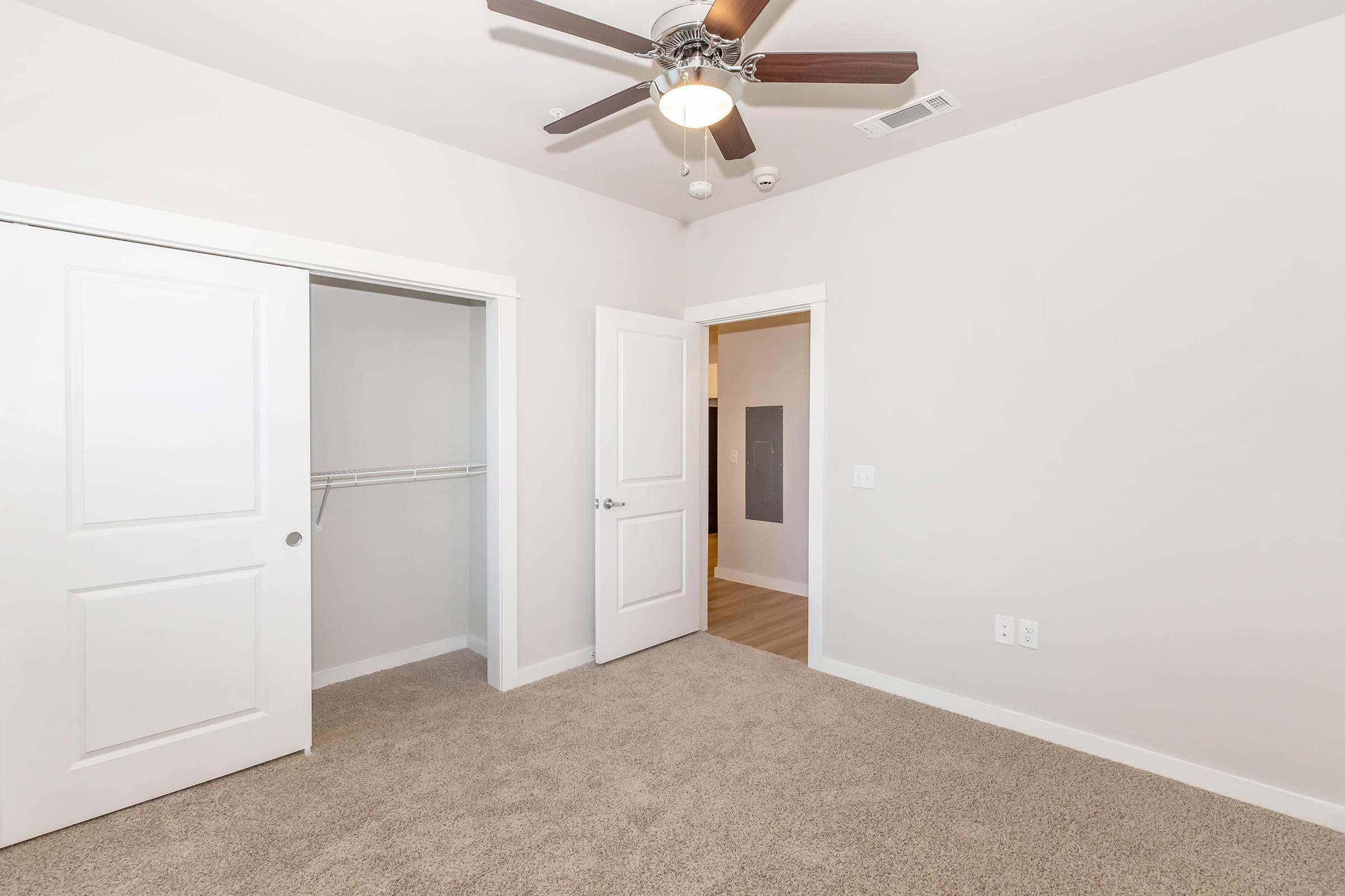 A well-lit room with light beige carpet, featuring a ceiling fan, white walls, and two doors. One door leads to a closet with a hanging rod, while the other opens to an adjacent space. The room has a modern, clean appearance, making it suitable for various uses, such as a bedroom or office.