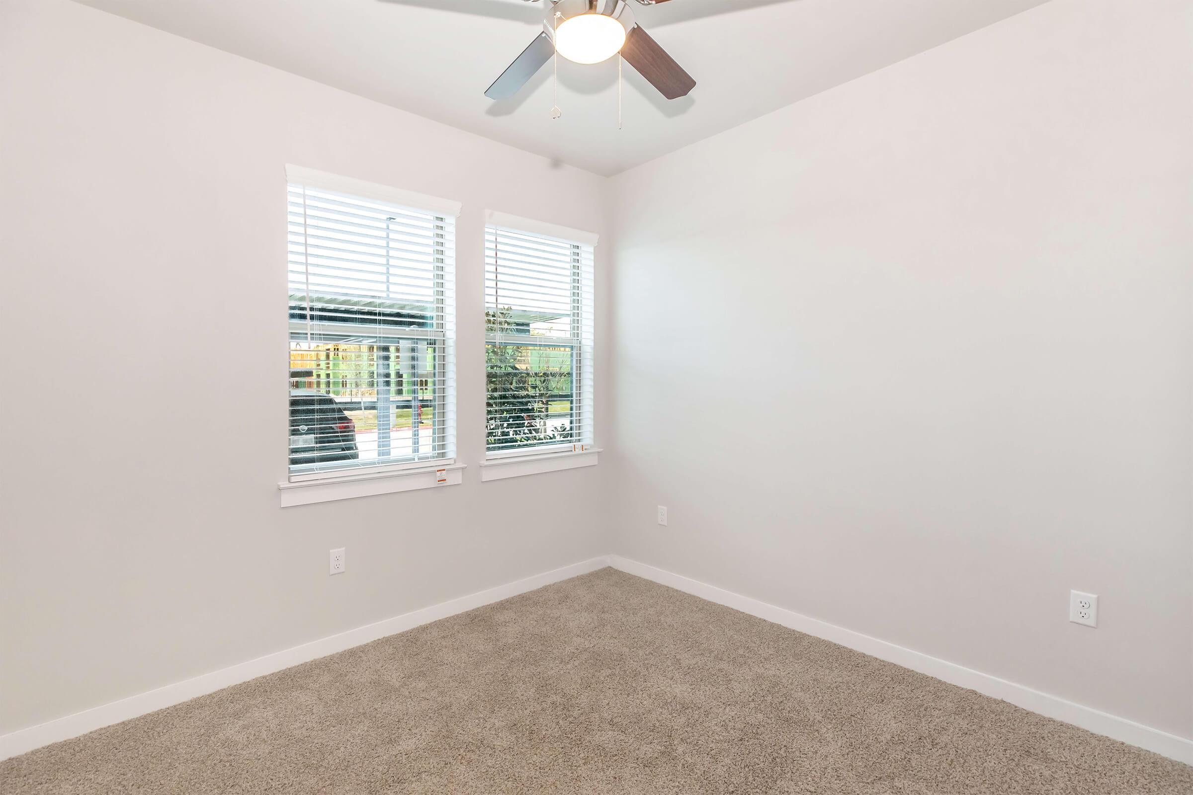 A well-lit, empty room with light-colored walls and a plush carpet. There are two windows with white blinds letting in natural light, and a ceiling fan with wooden blades. No furniture or decor is present, creating a spacious and minimalistic atmosphere.