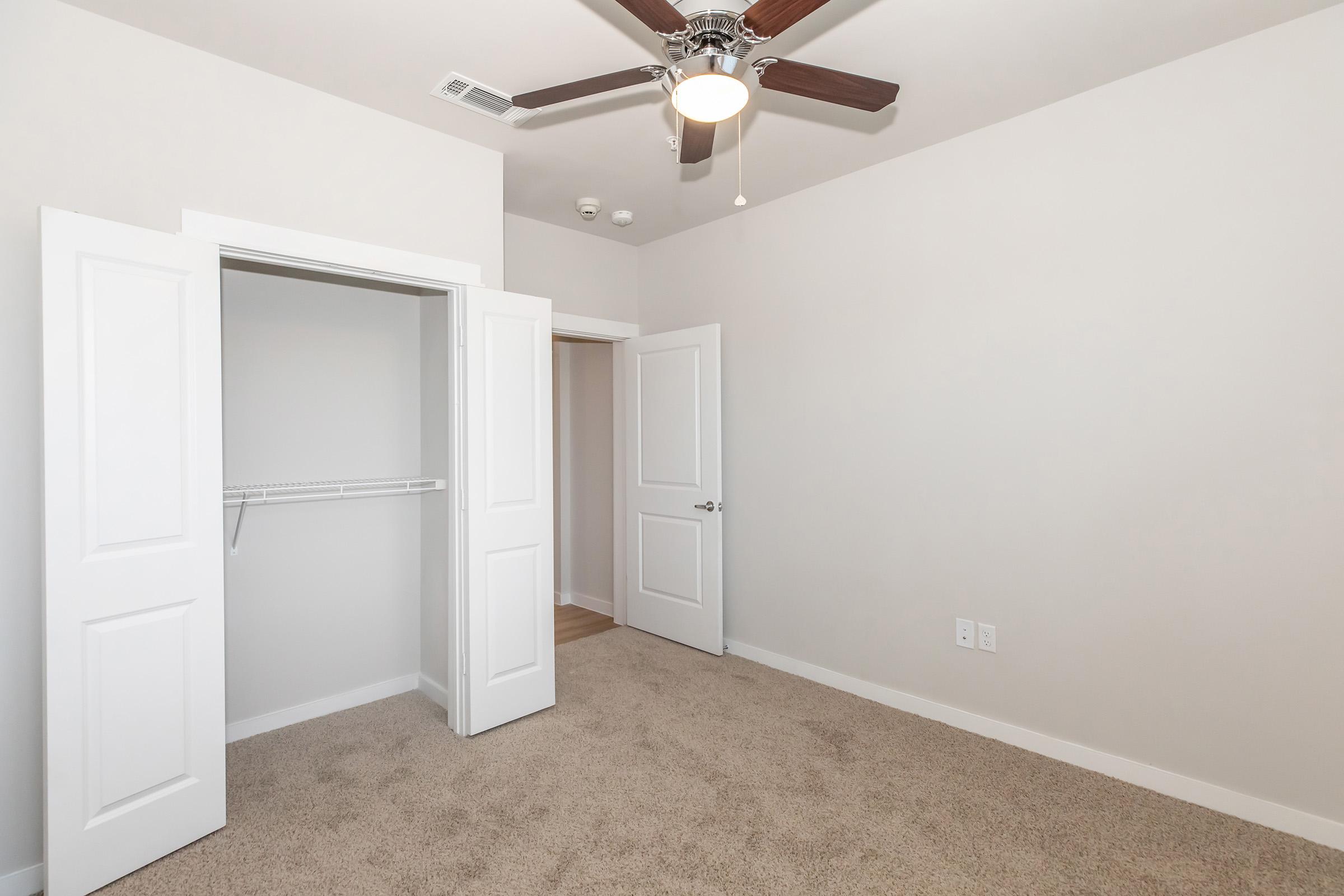 A light and airy room with beige carpeting, featuring a ceiling fan and two white doors. One door leads to a closet with a shelf, while the other opens to a hallway. The walls are painted a soft neutral color, enhancing the spacious feel of the room.