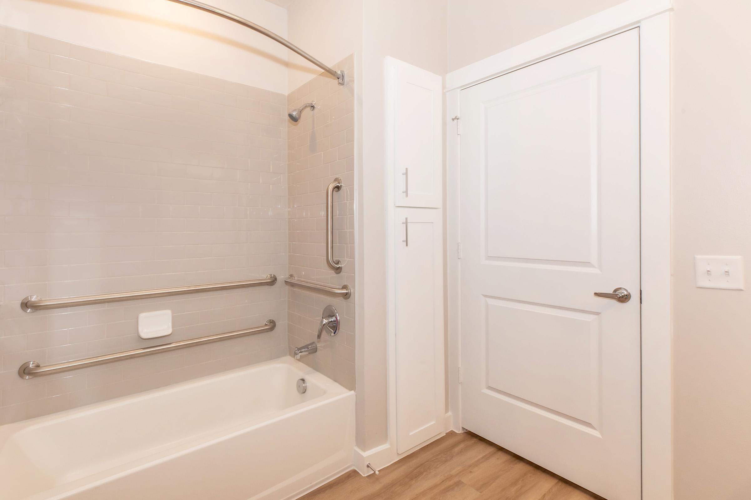 A clean, modern bathroom featuring a bathtub with grab bars, a tiled shower area, and a door leading to another room. The walls are painted in a light color, and there is a storage cabinet beside the bathtub. The floor has a wood-like finish, adding warmth to the space.