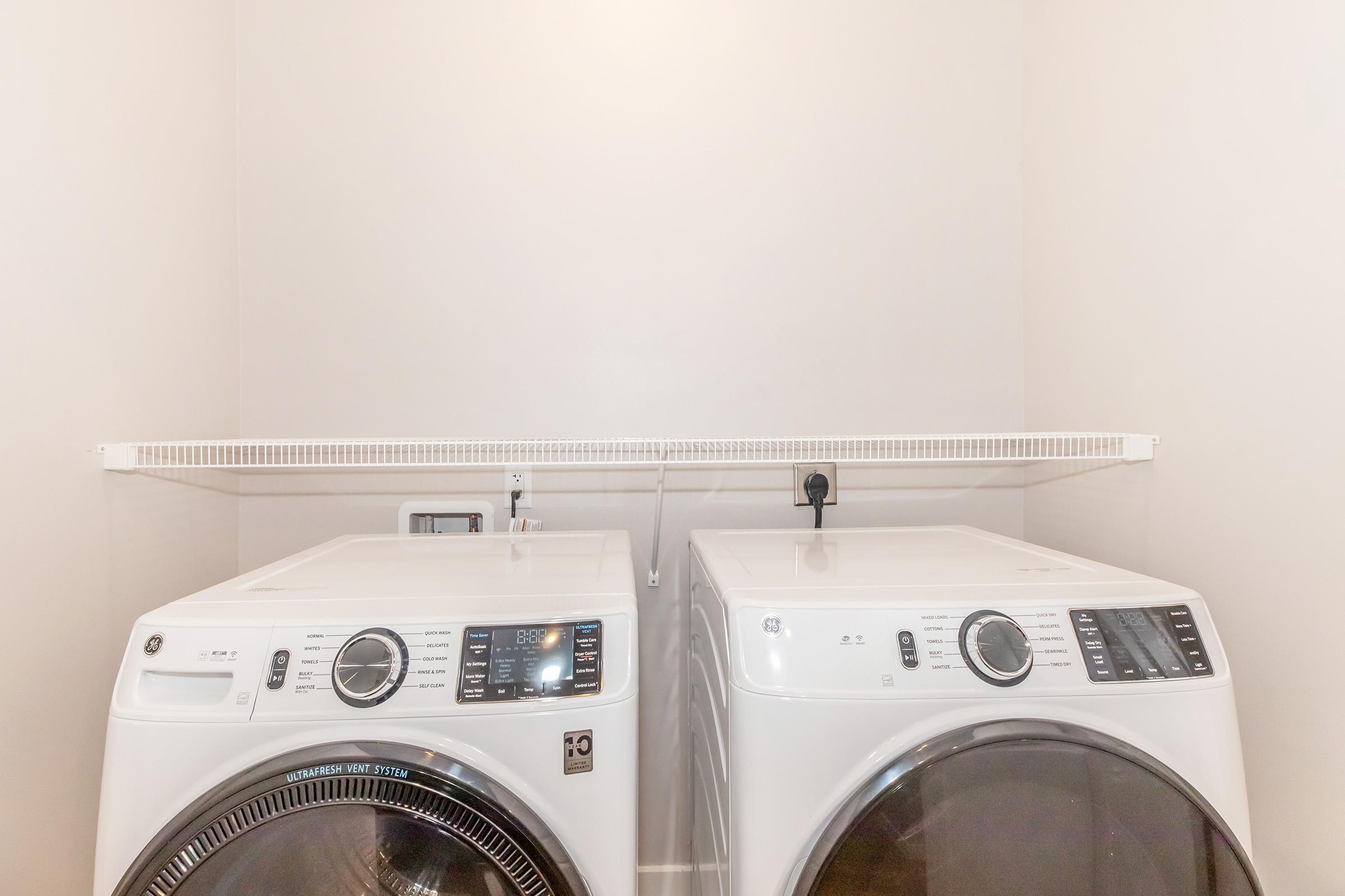 A clean laundry room featuring two white washing machines side by side on a light-colored wall. A shelf above provides storage space, and there's a power outlet visible on the wall. The overall setting is bright and organized, perfect for laundry tasks.