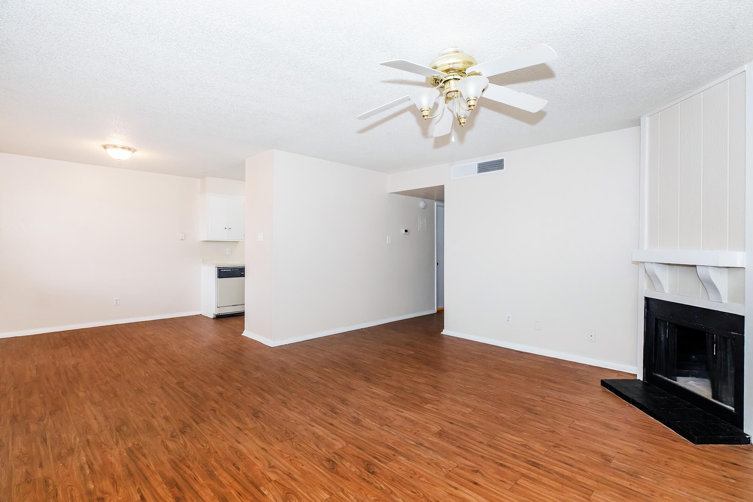Spacious living room with hardwood floors, ceiling fan, and fireplace. Light-colored walls and open layout leading to a kitchen. Natural light entering from multiple angles, creating a welcoming atmosphere.