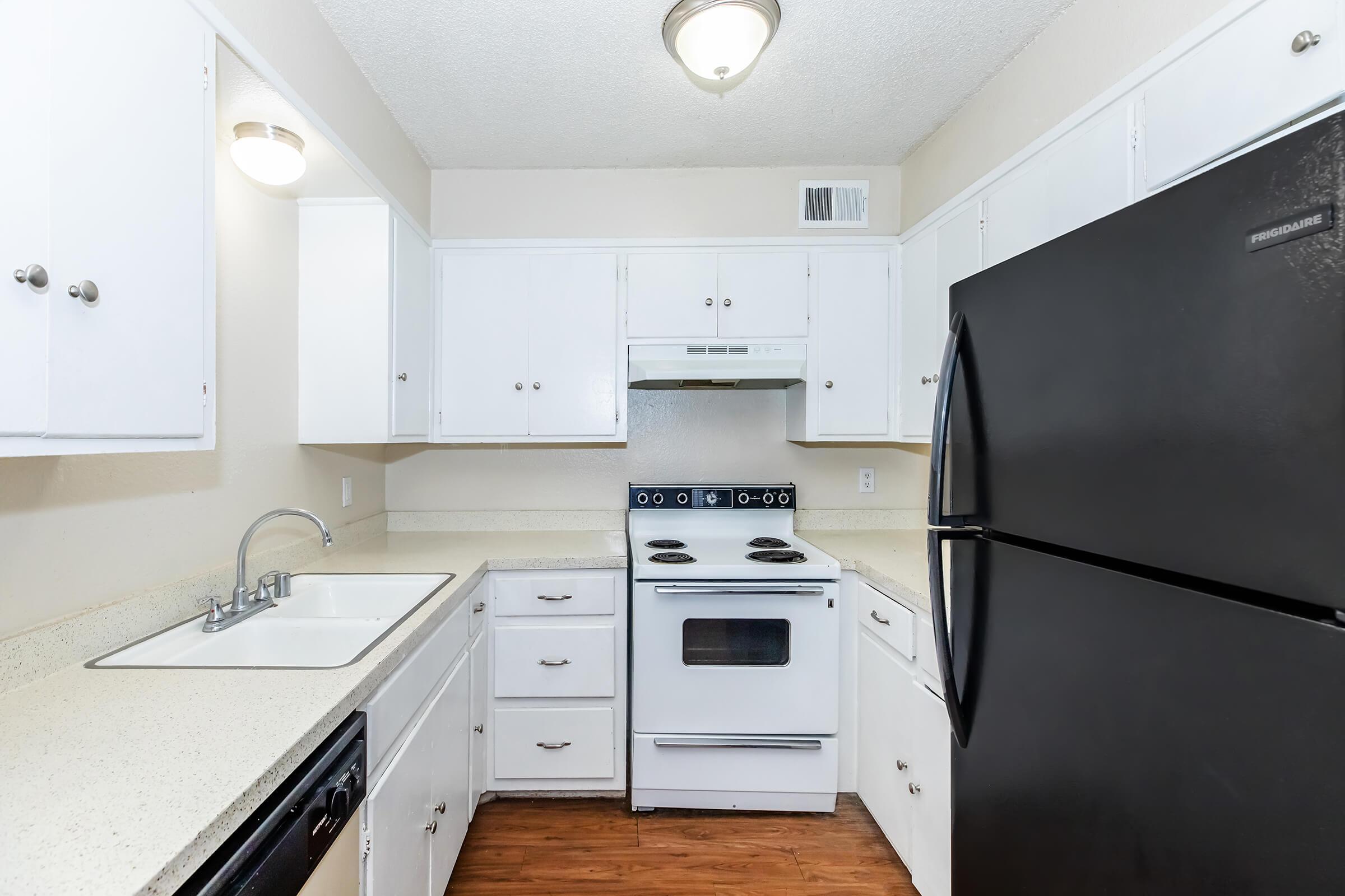 A small kitchen featuring white cabinetry, a double sink, a black refrigerator, and a white stove with an oven. The space has light-colored walls and wood-style flooring, creating a bright and functional cooking area.