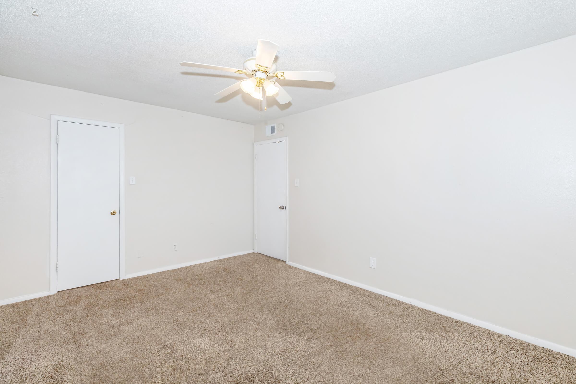 Empty room with light beige walls and cream-colored carpet. A ceiling fan with three blades is mounted in the center. There is a closed white door on the left and a second closed door on the right. The room appears spacious and well-lit, with no furniture present.
