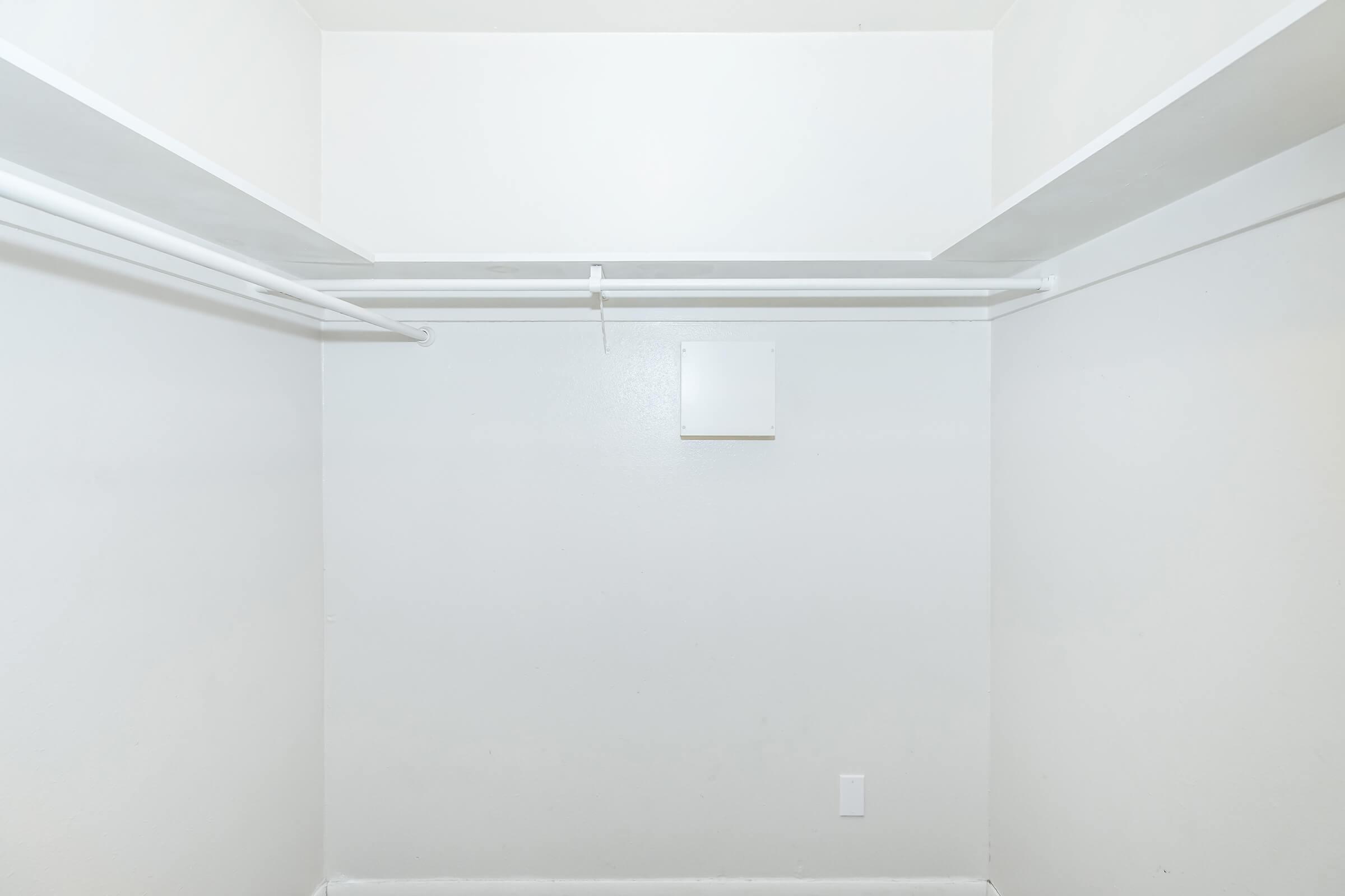 A spacious, empty walk-in closet with white walls and shelving. There are two horizontal rods for hanging clothes on either side, and a small vent or access panel on the back wall. The floor is bare, suggesting the closet is unused or waiting to be organized.